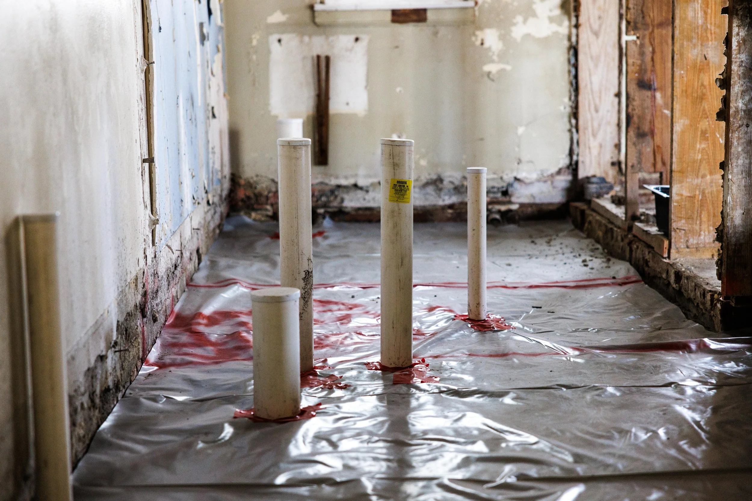 Inside a room under renovation, several white plumbing pipes protrude through plastic sheeting on the floor, with exposed walls and wooden framing in the background.