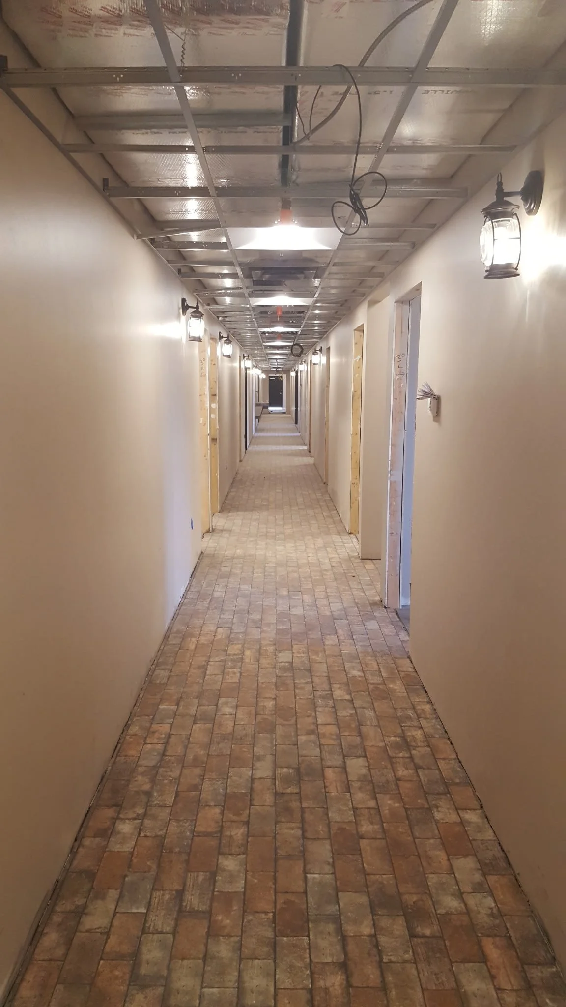 Long hallway under construction with brick flooring, beige walls, and unfinished doorways. Overhead lighting and wall-mounted lantern-style lights are present, with exposed ceiling wiring and ductwork.
