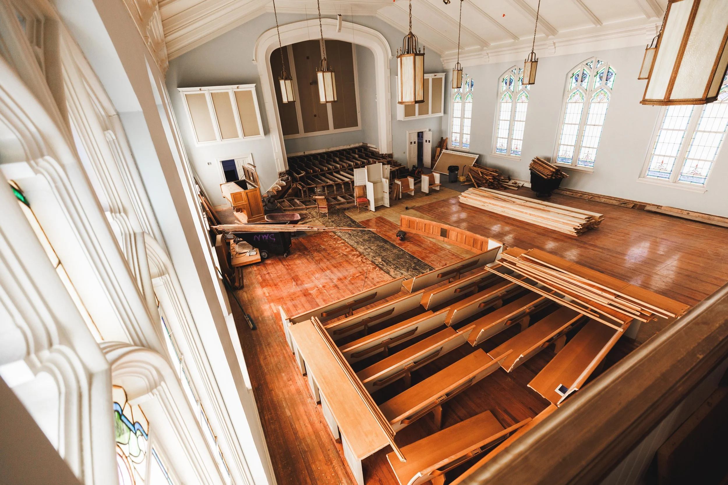 Interior of a church under renovation, with wooden pews, a stage with a rug, stained glass windows, and construction materials scattered around.