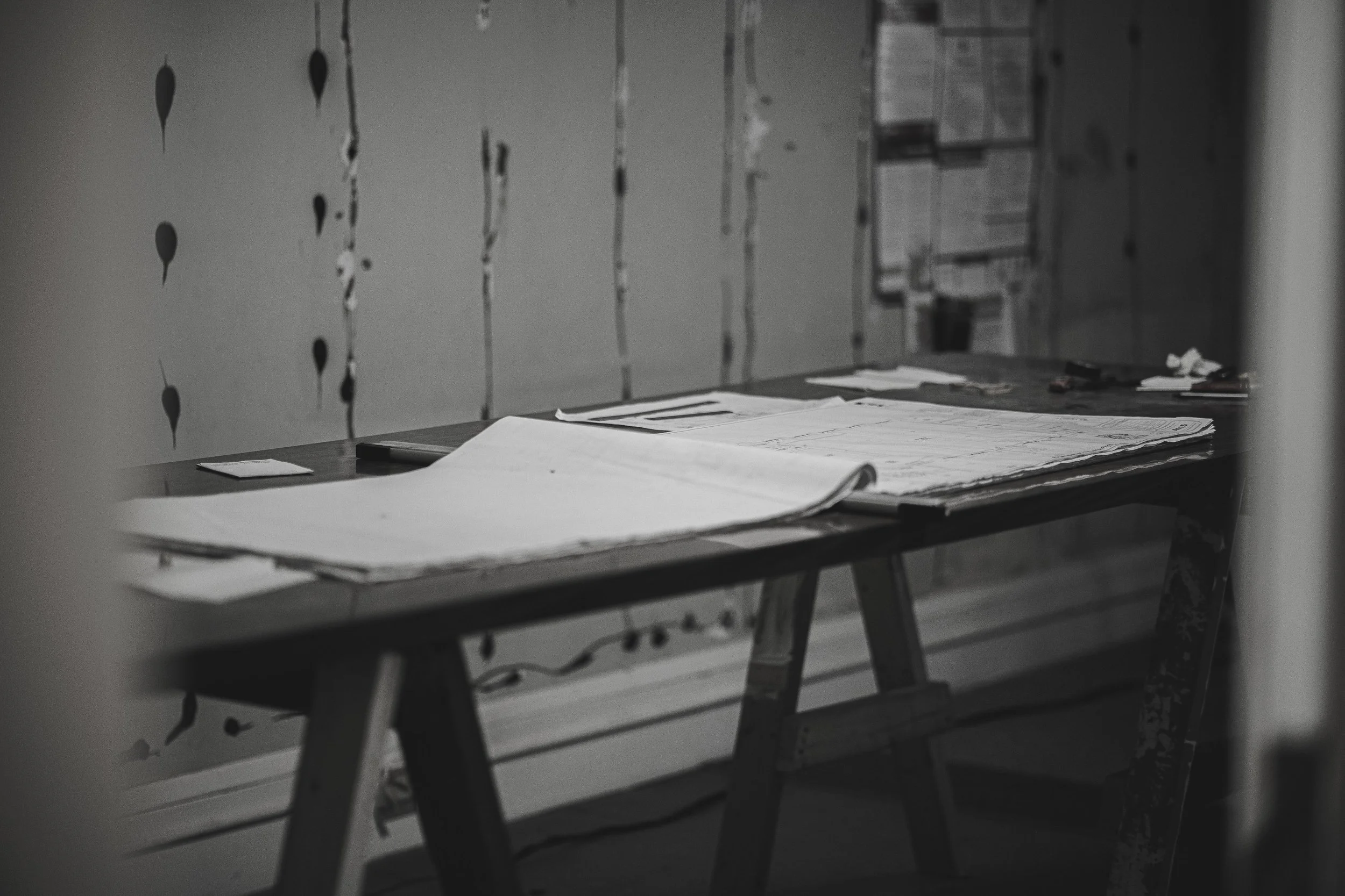 A desk with blueprints and papers on it in an unfinished room with a wall featuring painted black irregular shapes.
