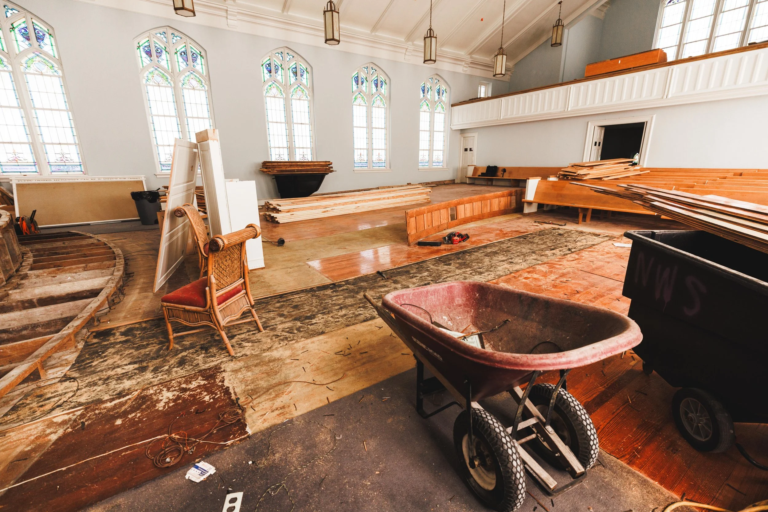 Church interior under renovation with construction tools, pliable wood sheets, a wheelbarrow, and scattered construction materials, with stained glass windows and hanging lights.