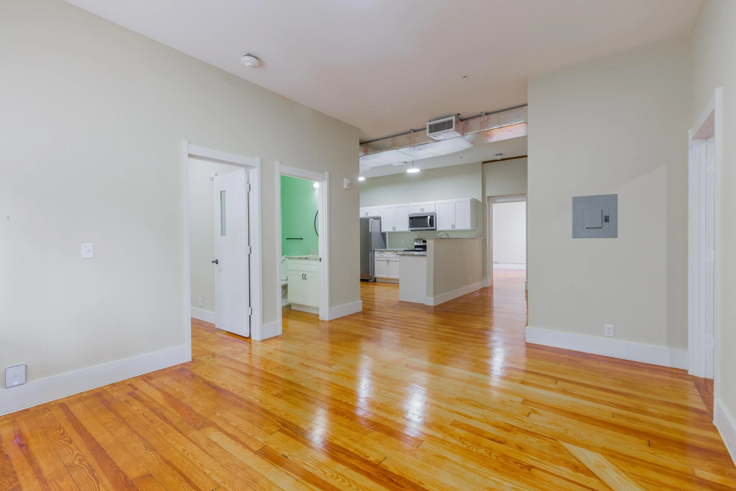 Empty apartment with hardwood floors, white walls, and an open kitchen with white cabinets and stainless steel appliances.