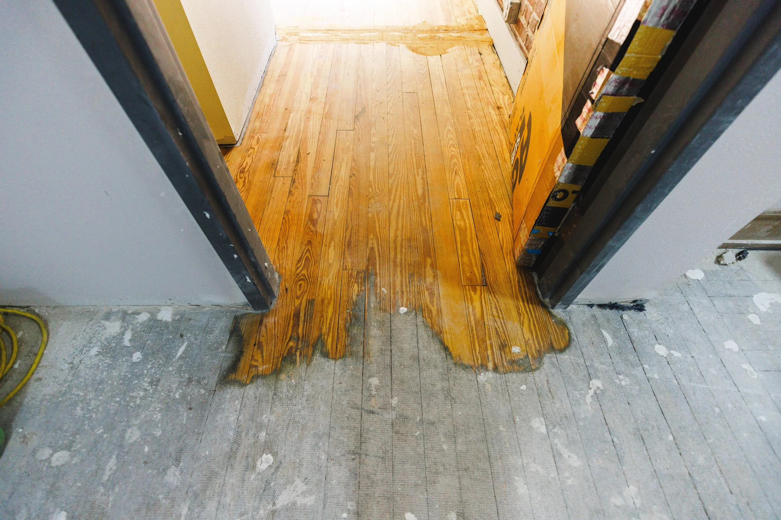 A doorway with a partially finished wooden floor transition from concrete to hardwood flooring. The hardwood has a glossy finish, while the concrete is rough and unfinished. Construction supplies and tools are visible nearby.