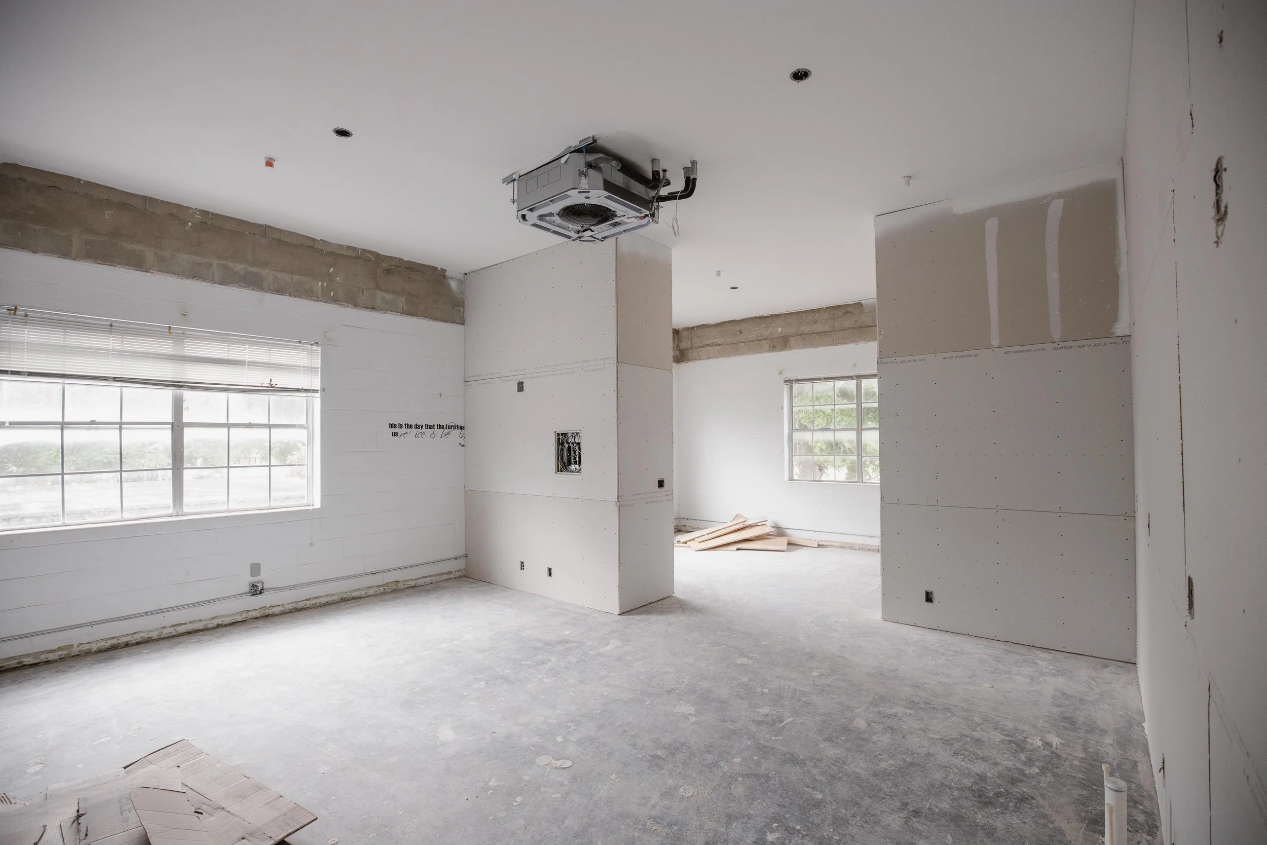 Empty room under construction with unfinished drywall, exposed window frames, and a ceiling air conditioning unit. Piles of wood and construction materials are on the floor.
