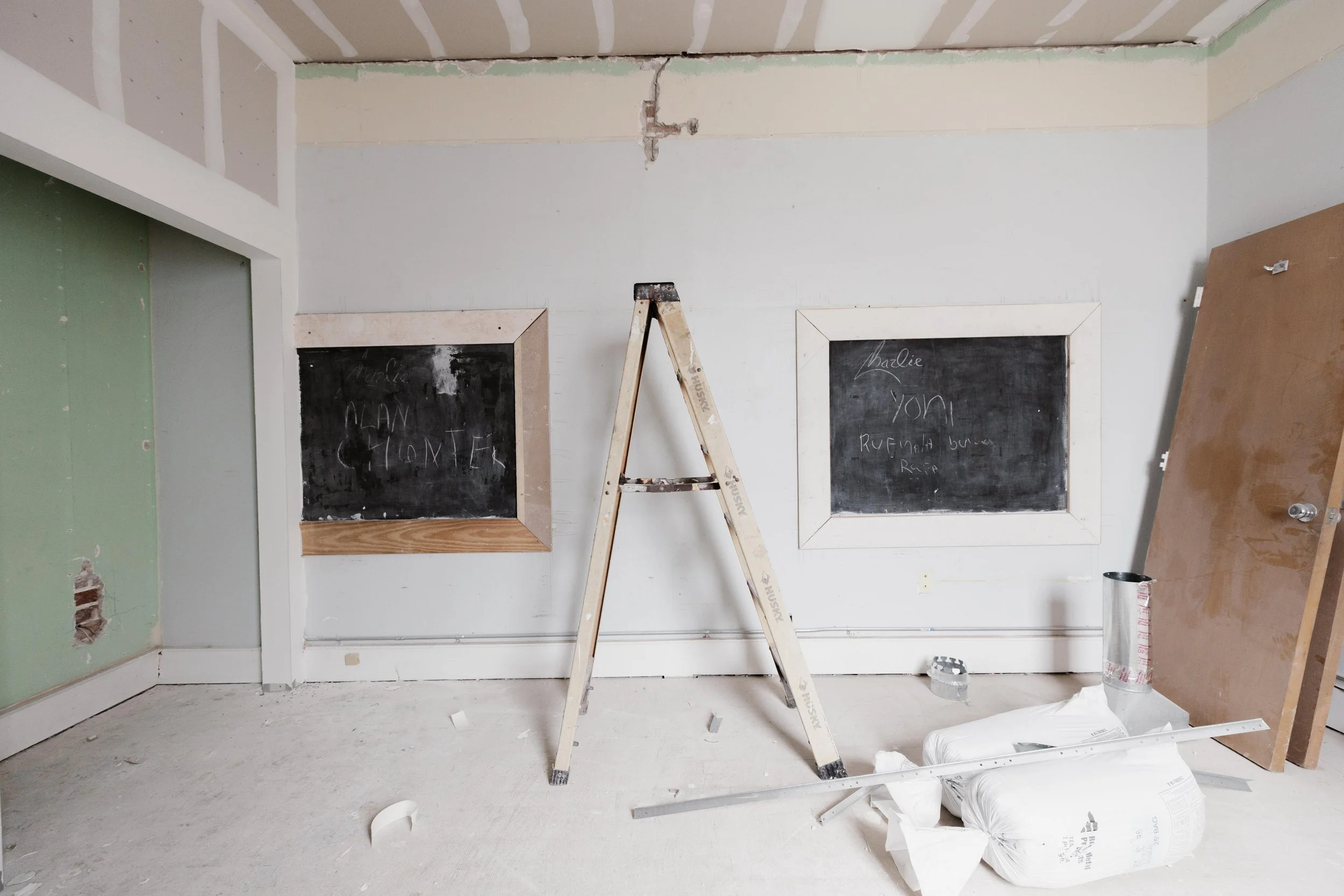 Construction site of an interior wall with two chalkboards, a step ladder, construction materials, and tools.