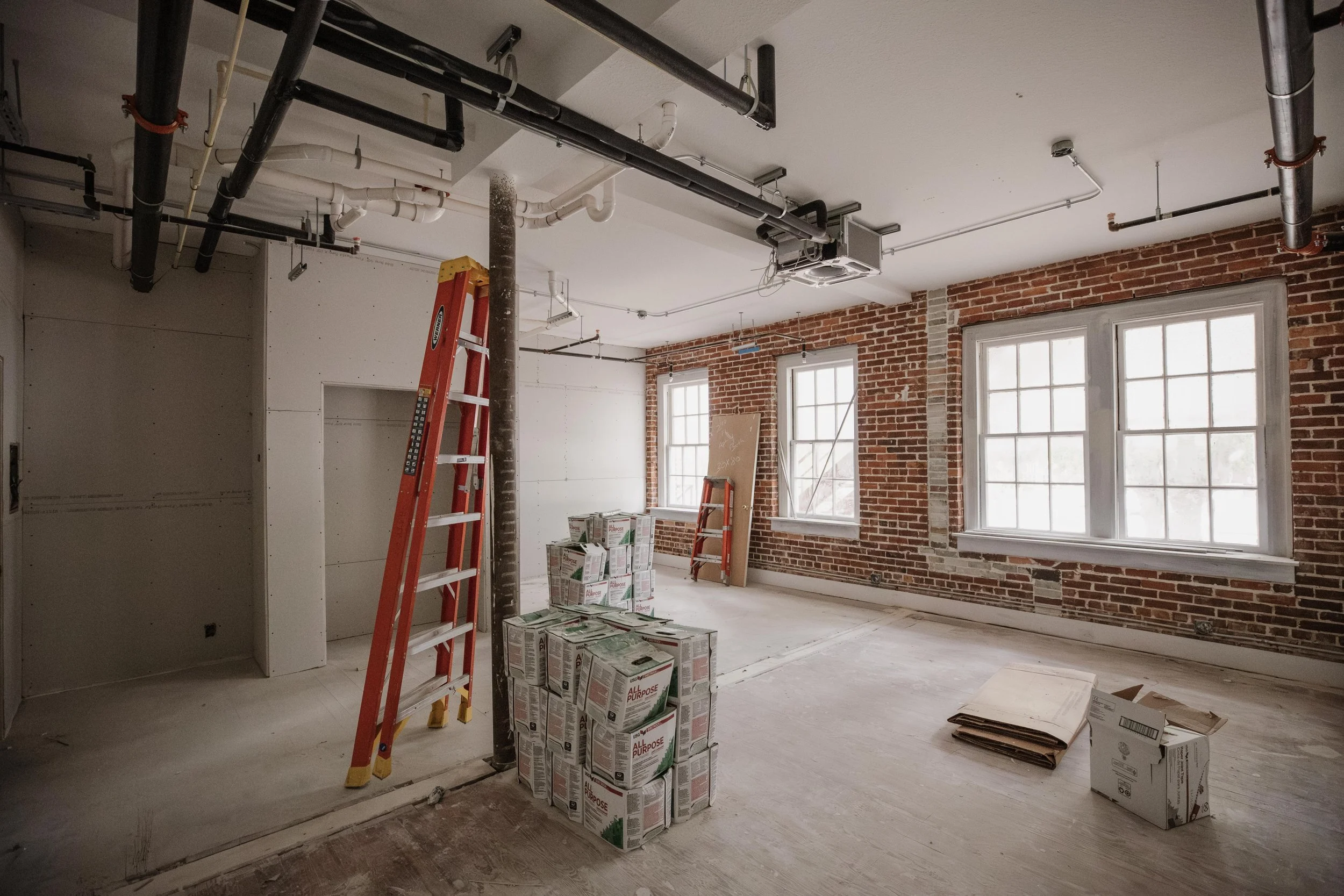 Interior of a room under construction with brick walls, three large windows, ladders, boxes of building materials, and exposed pipes on the ceiling.