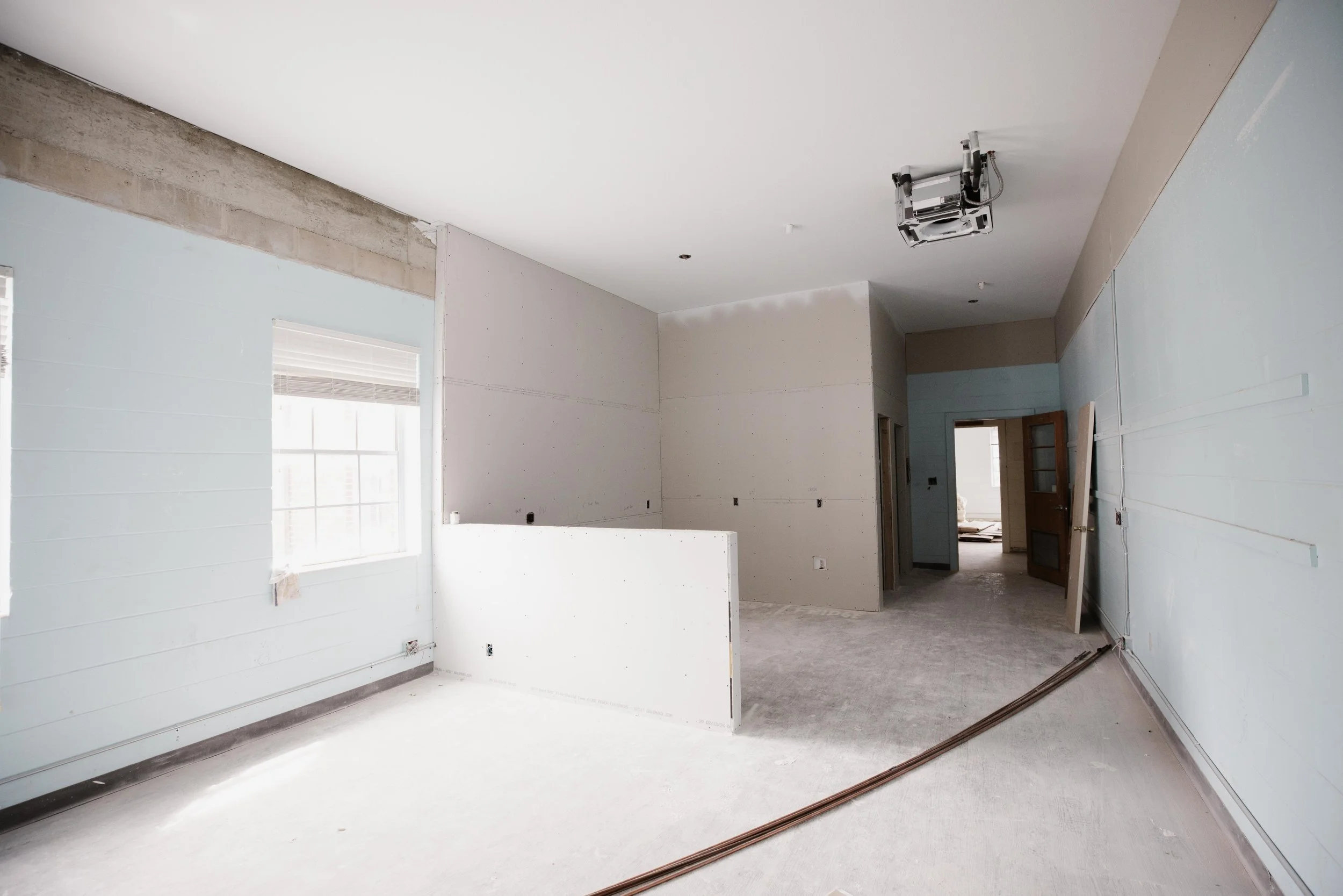 Interior of a house under construction with unfinished walls, electrical wiring, and a mounted ceiling fixture, natural light coming through windows.