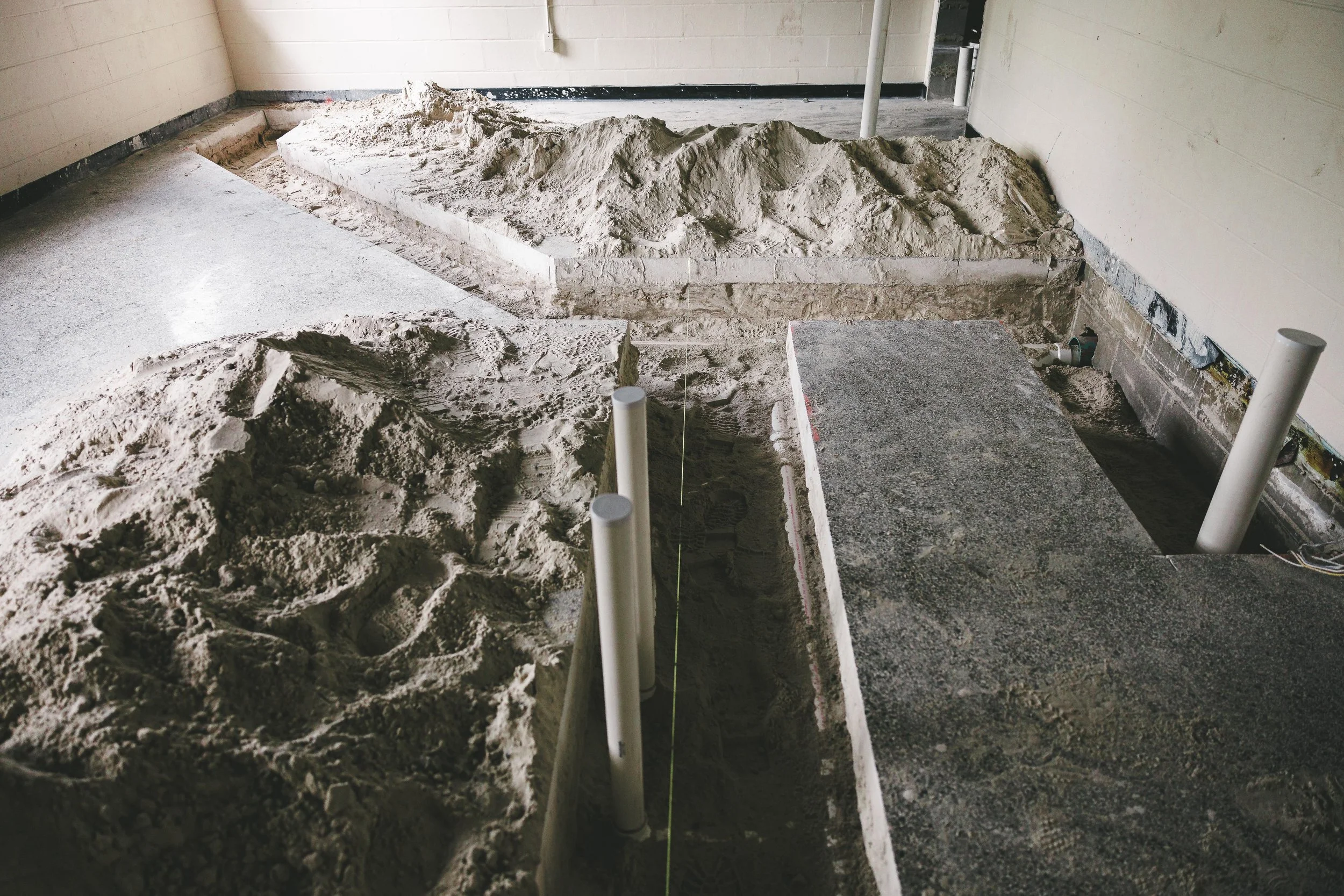 Construction site showing sand, concrete, and plumbing pipes prepared for building installation.