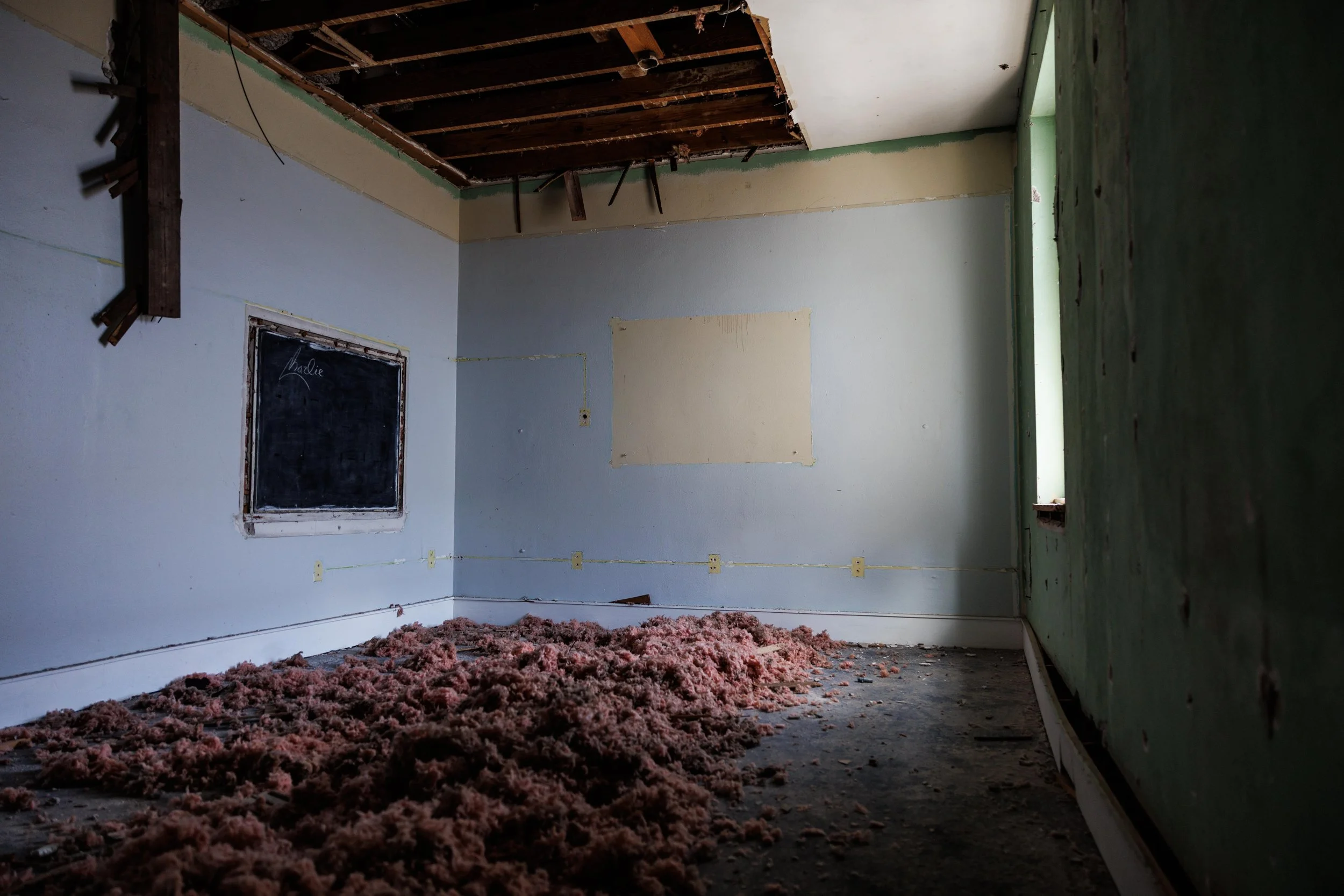 An abandoned room under renovation with drywall and insulation exposed, a window on the right side, and pink insulation scattered on the dirty floor