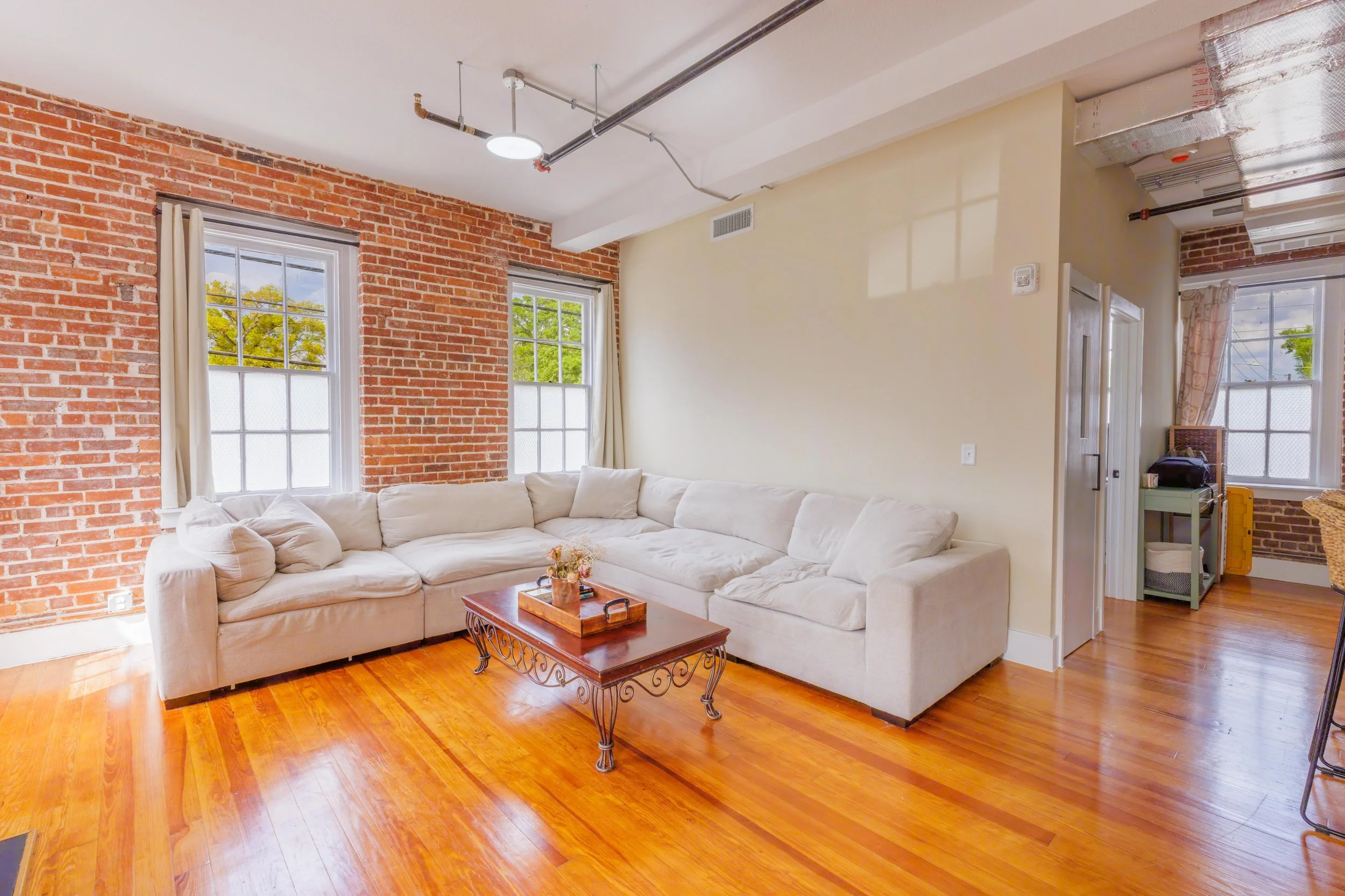 Living room with a beige sectional sofa, wooden coffee table, exposed brick wall, and hardwood floors. There are three windows with white curtains and a ceiling light fixture.