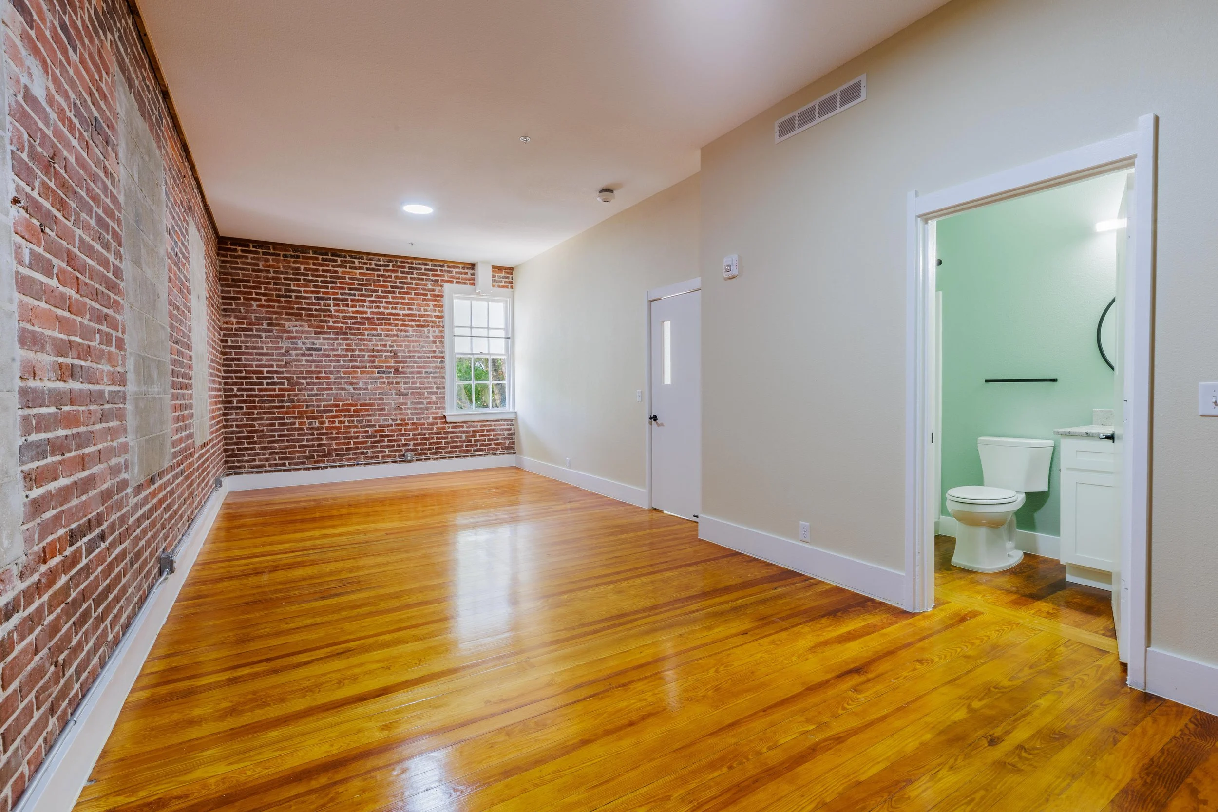 Empty living room with hardwood floors, exposed brick wall, window, and adjacent small bathroom with toilet and sink.