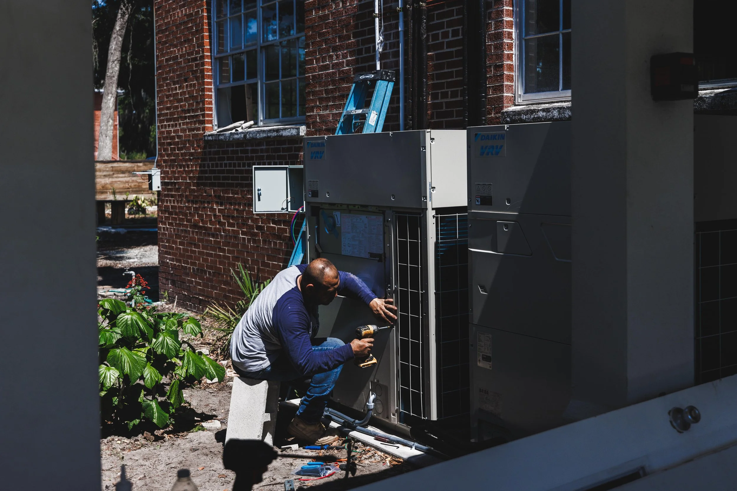 A man working on an outdoor HVAC unit next to a brick building, using a power drill on the unit.