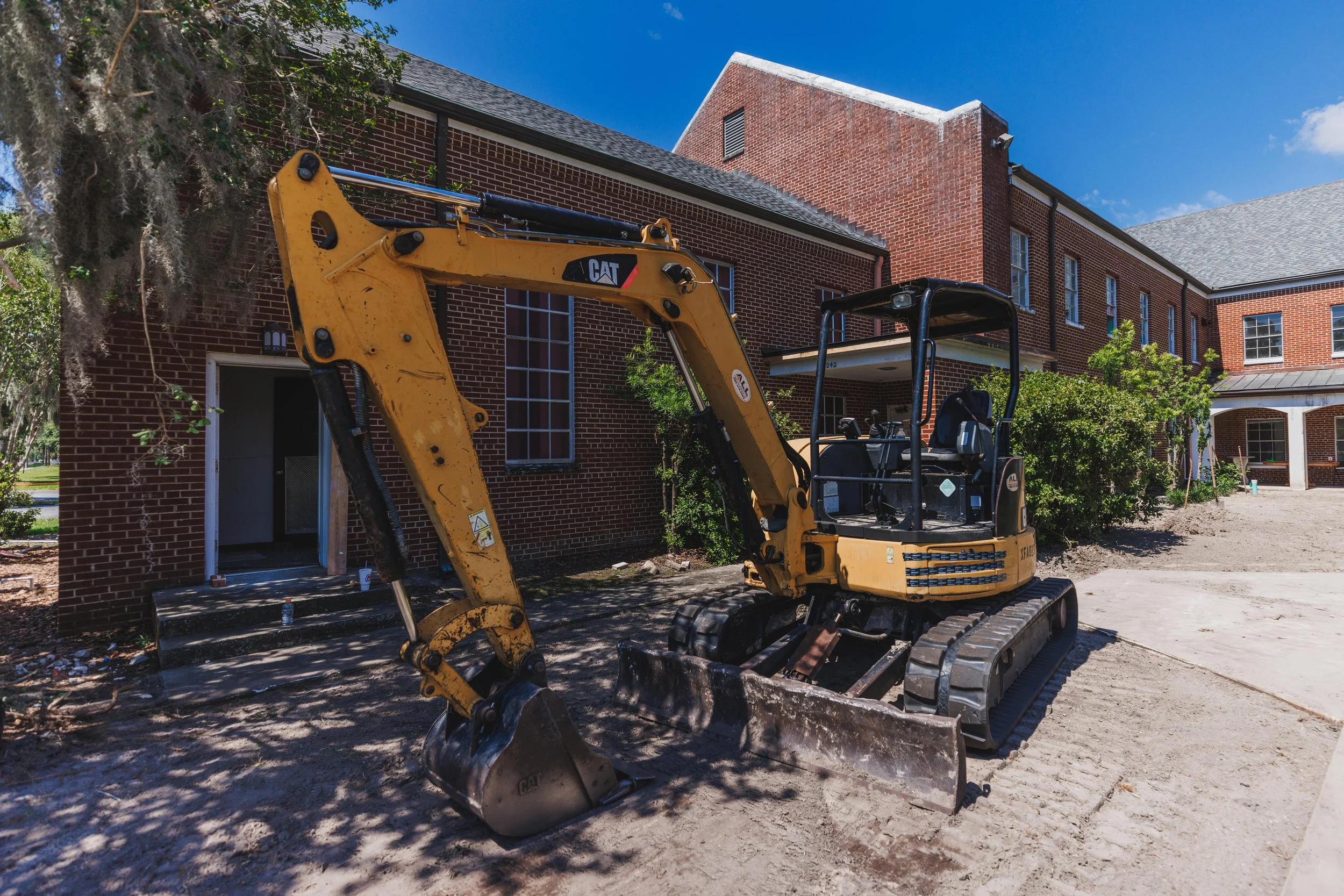 A yellow Caterpillar mini excavator parked in front of a brick building under a clear blue sky.
