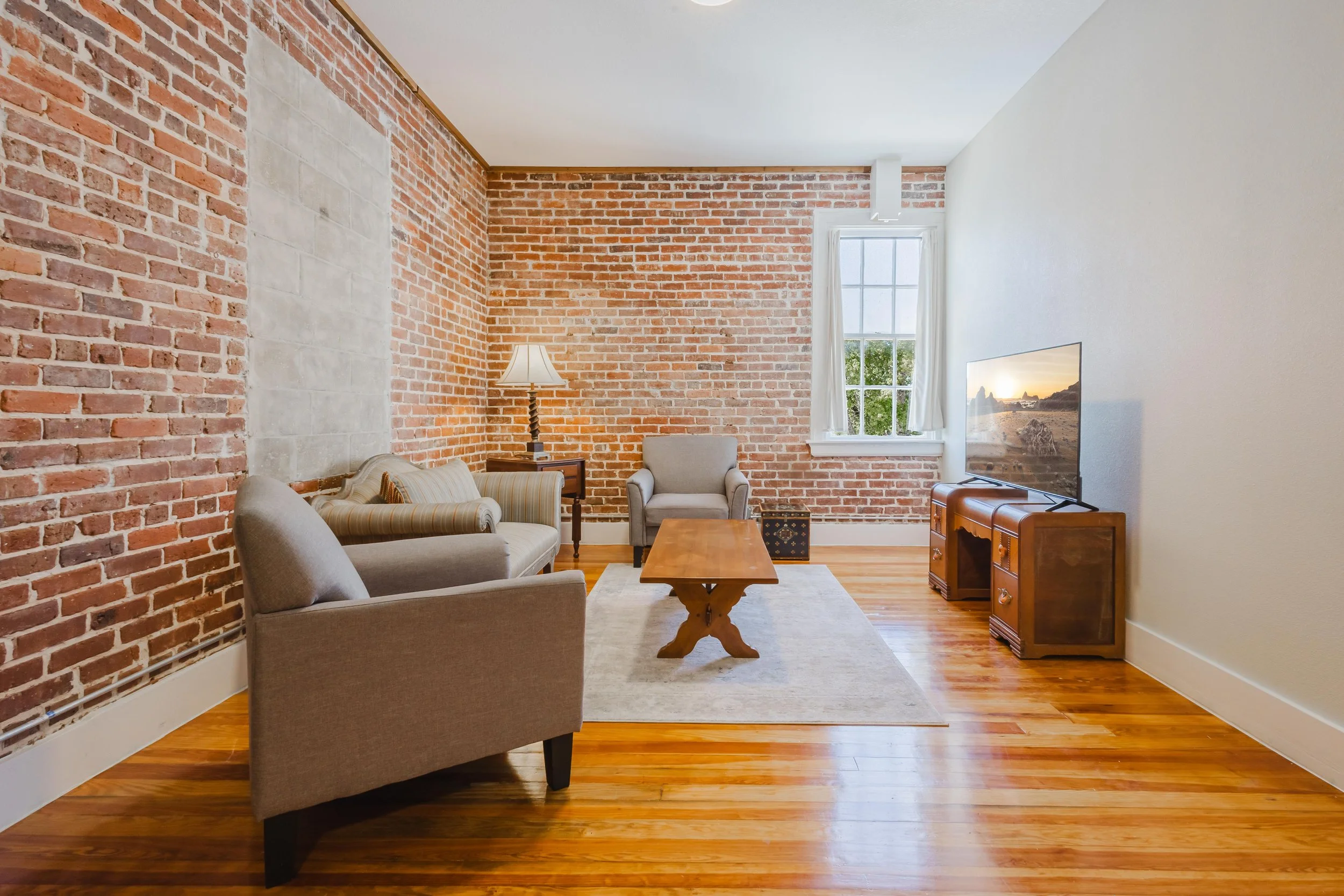 Living room with exposed brick wall, hardwood floors, beige and gray furniture, a wooden coffee table, a TV on a wooden stand, window with white curtains, and a lamp on a side table.