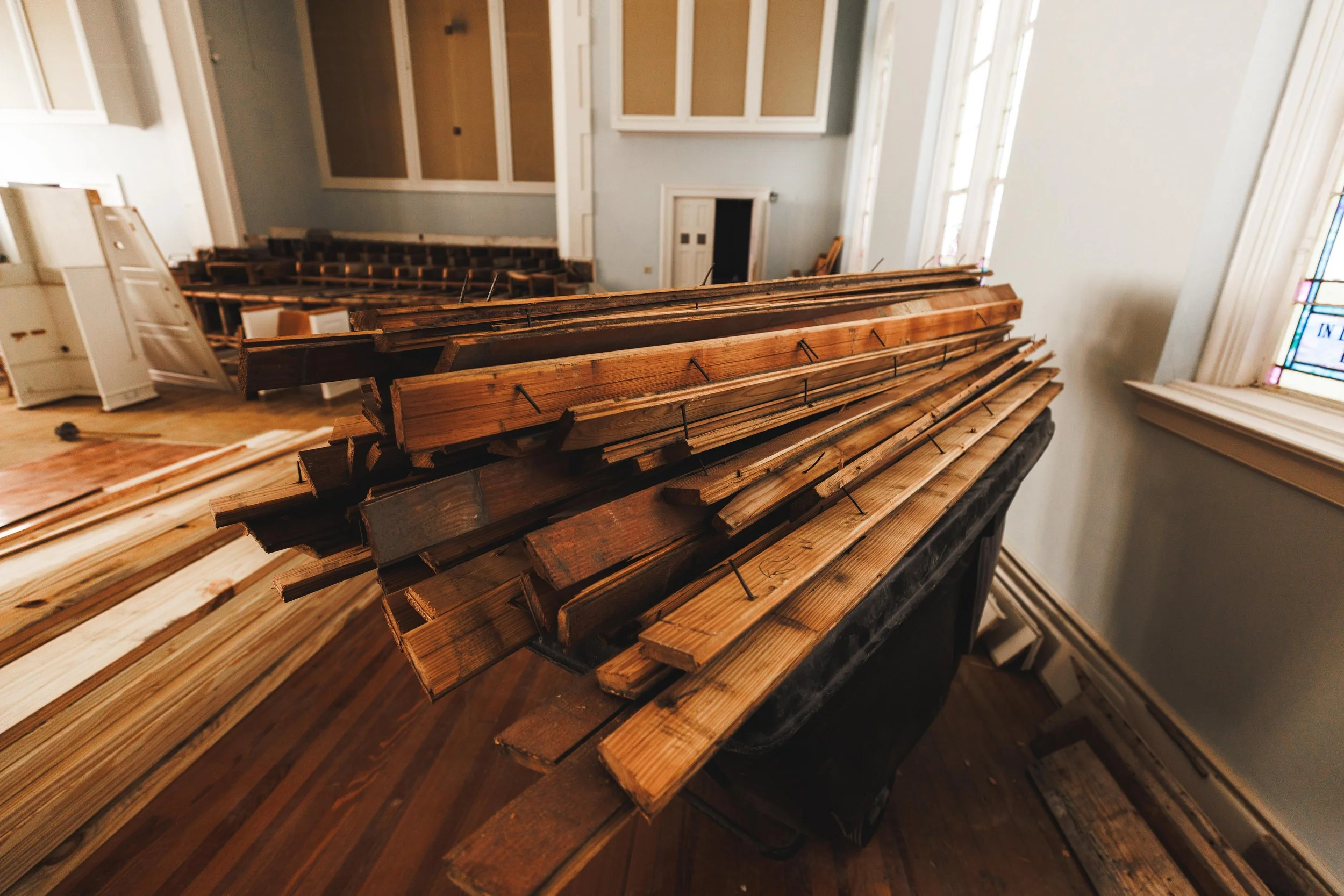 Piles of wooden floorboards stacked on a table inside a church or hall under renovation, with some windows and chairs in the background.