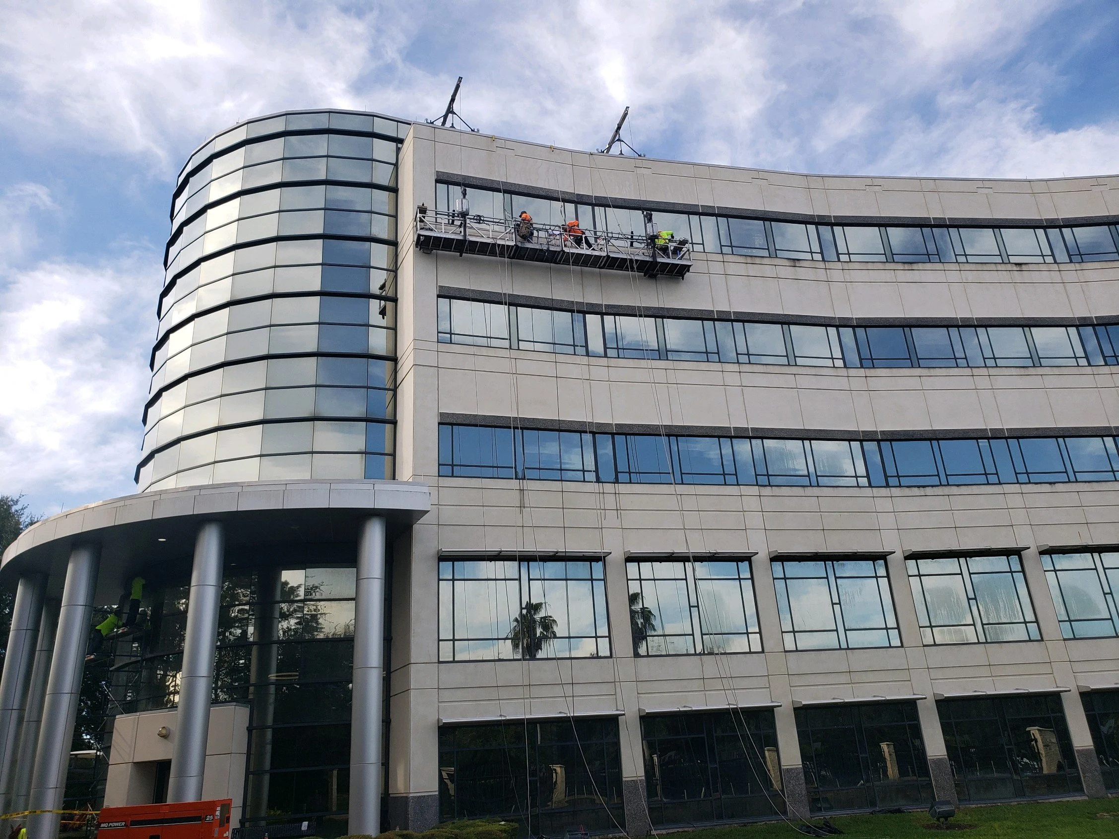 Workers on a suspended platform cleaning or maintaining the exterior windows of a modern multi-story office building with glass and concrete facade.
