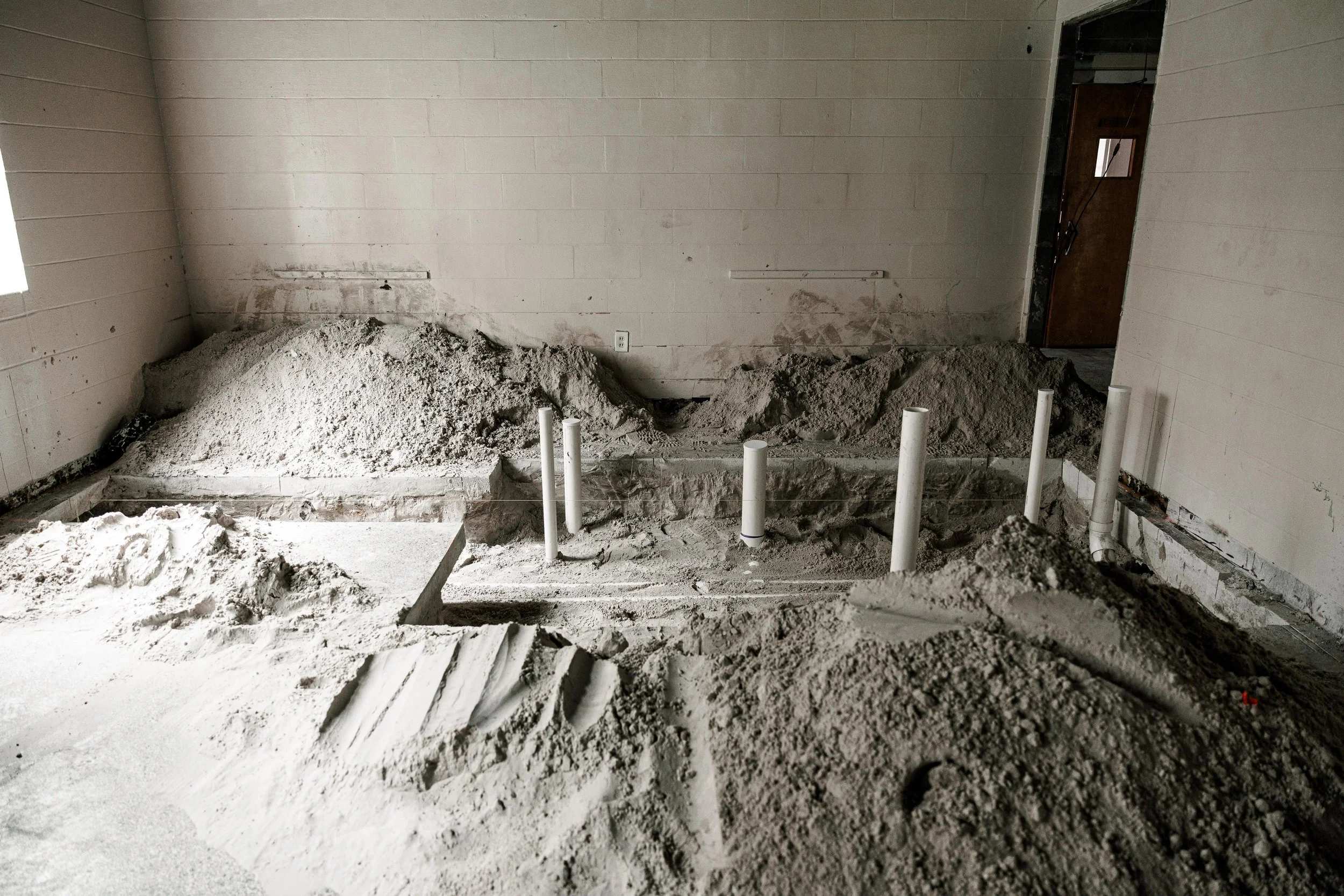 Room under construction with scattered sand and four PVC pipes protruding from the ground, with some piles of sand and an unfinished doorway in the background.