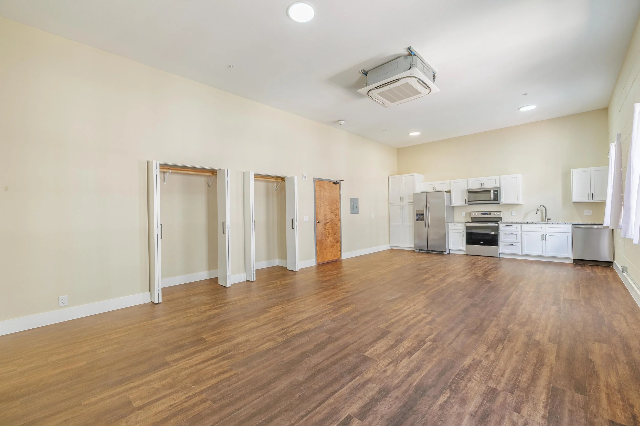 Empty room with hardwood floors, kitchen area with white cabinets, stainless steel appliances, and three open closet spaces with white folding doors.