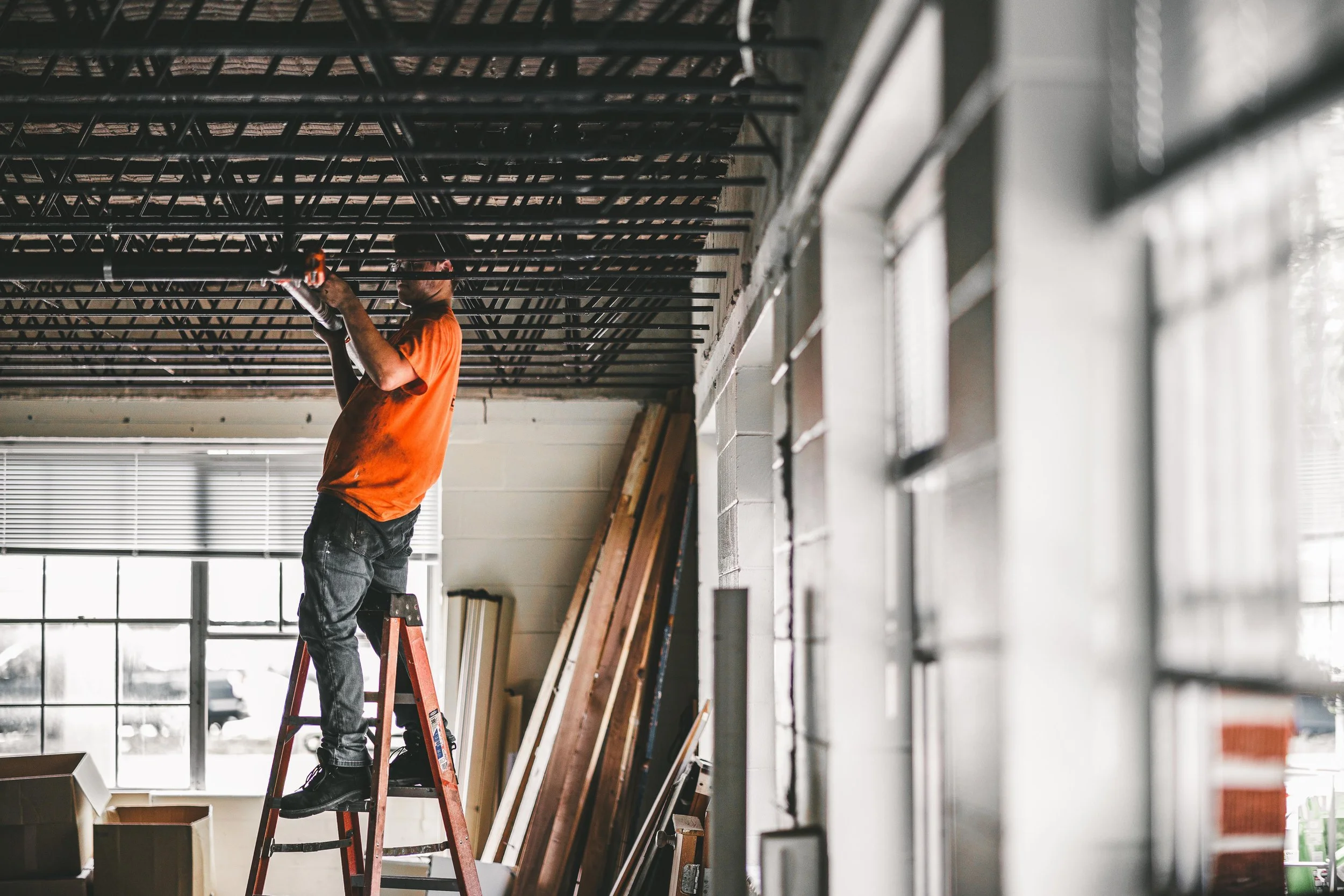 A person standing on a ladder working on a suspended ceiling structure in a room with large windows and tools, surrounded by construction materials.
