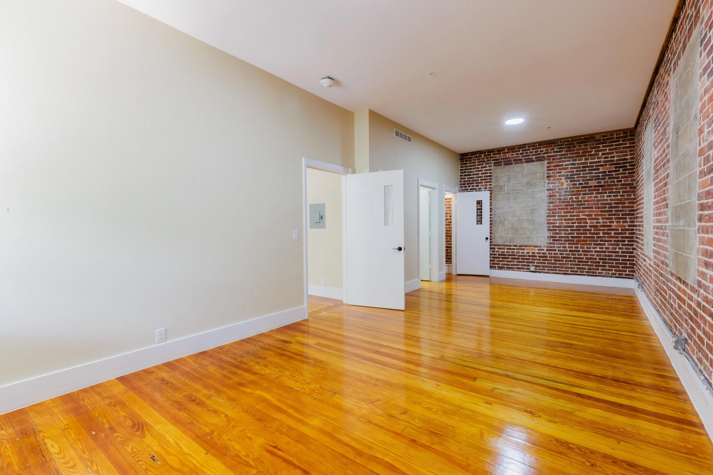 Empty room with polished wooden floors, cream-colored walls, and a textured brick wall with patches of different brick patterns.