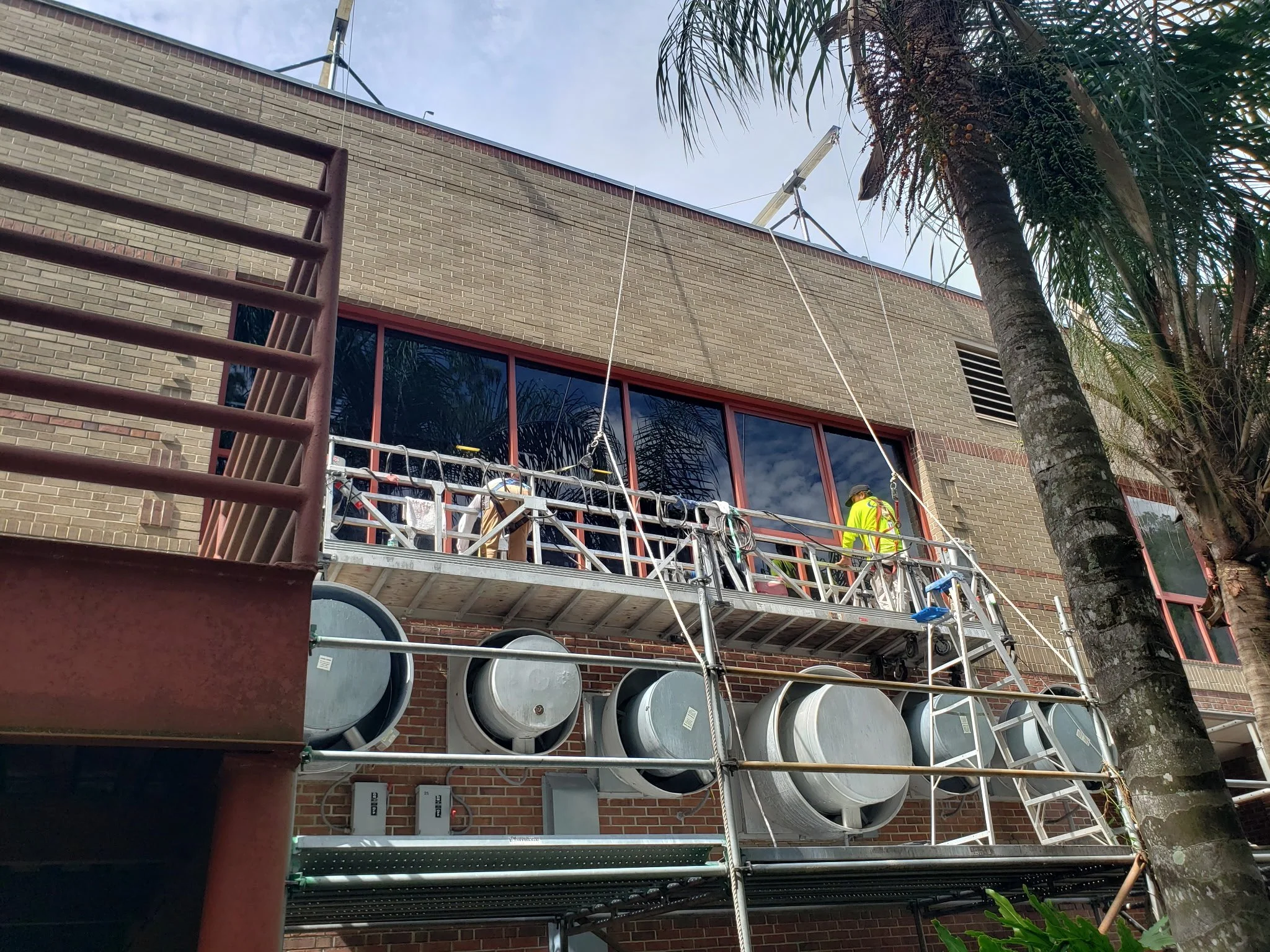 Construction workers on scaffolding cleaning or repairing the windows of a brick building, with large ventilation ducts and ventilation fans below.