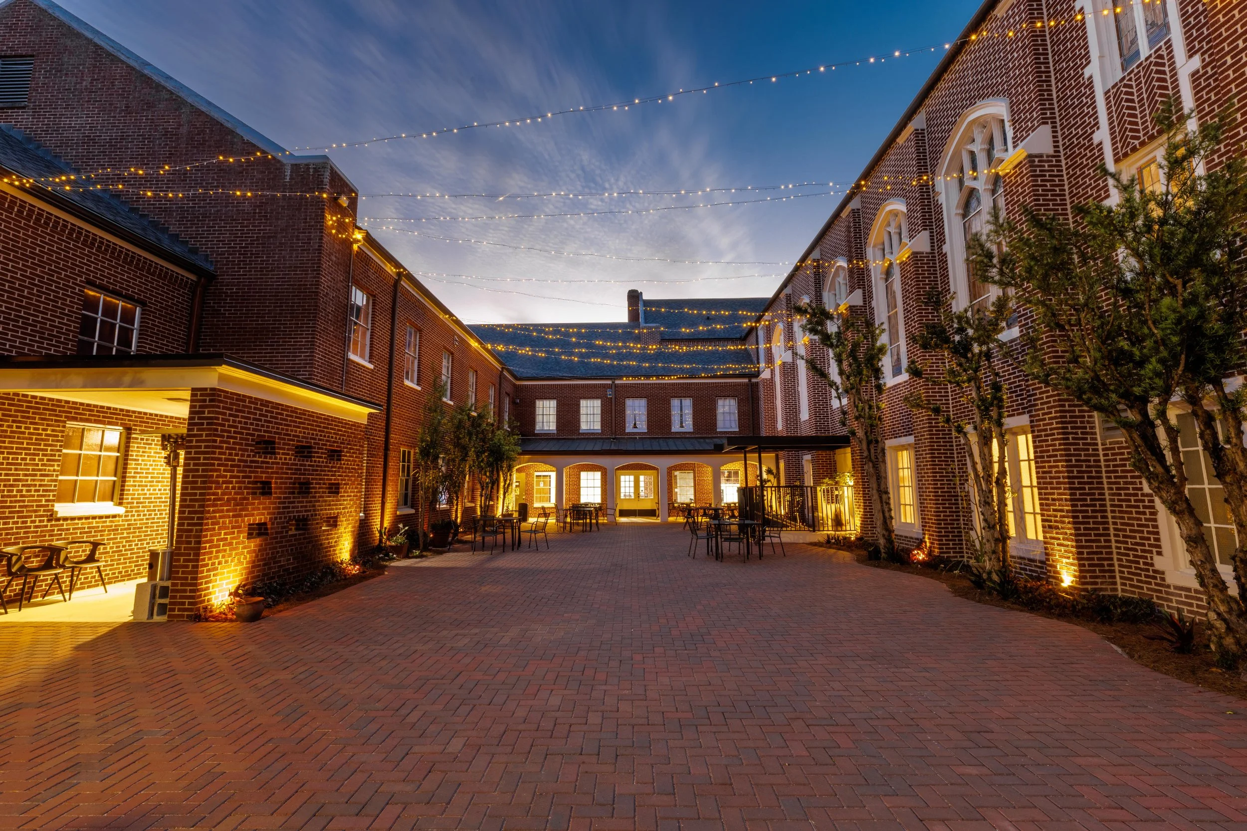 An outdoor courtyard illuminated with string lights, brick buildings surrounding the space, and trees with lights, during dusk.