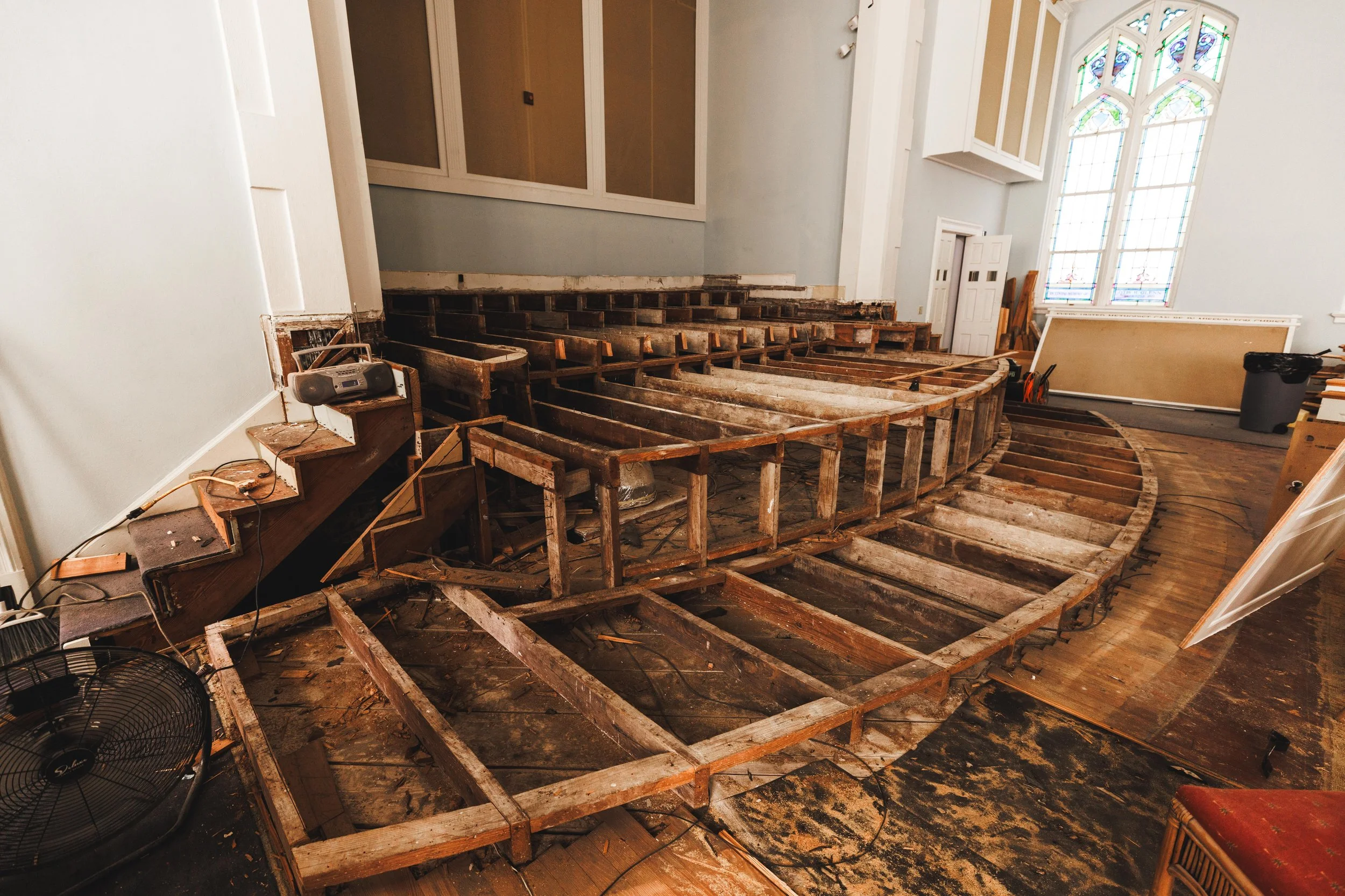 Interior of a church under renovation, showing wooden chair frames removed, exposing the curved floor underneath, with construction tools and debris on the floor.