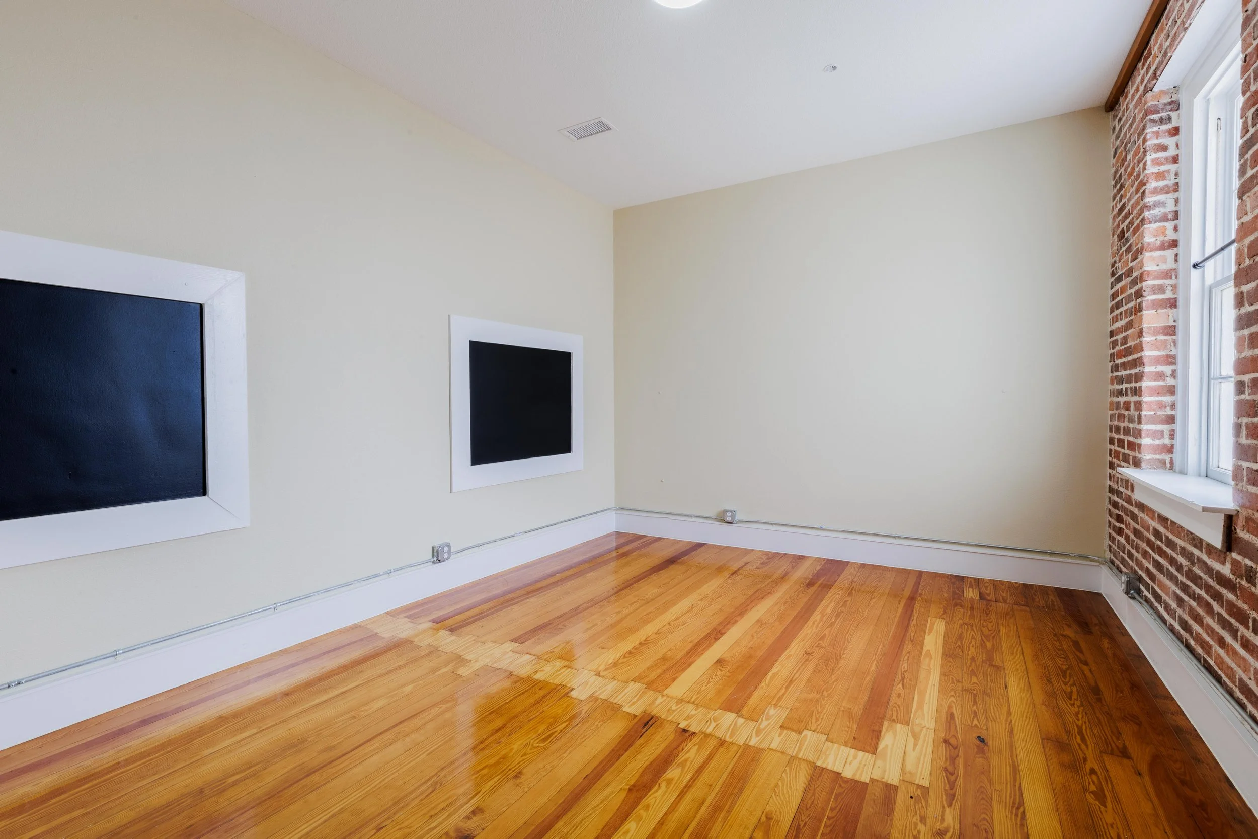 Empty room with hardwood floors, beige walls, exposed brick wall on the right, two blackboards on the left wall, and a window on the right wall.