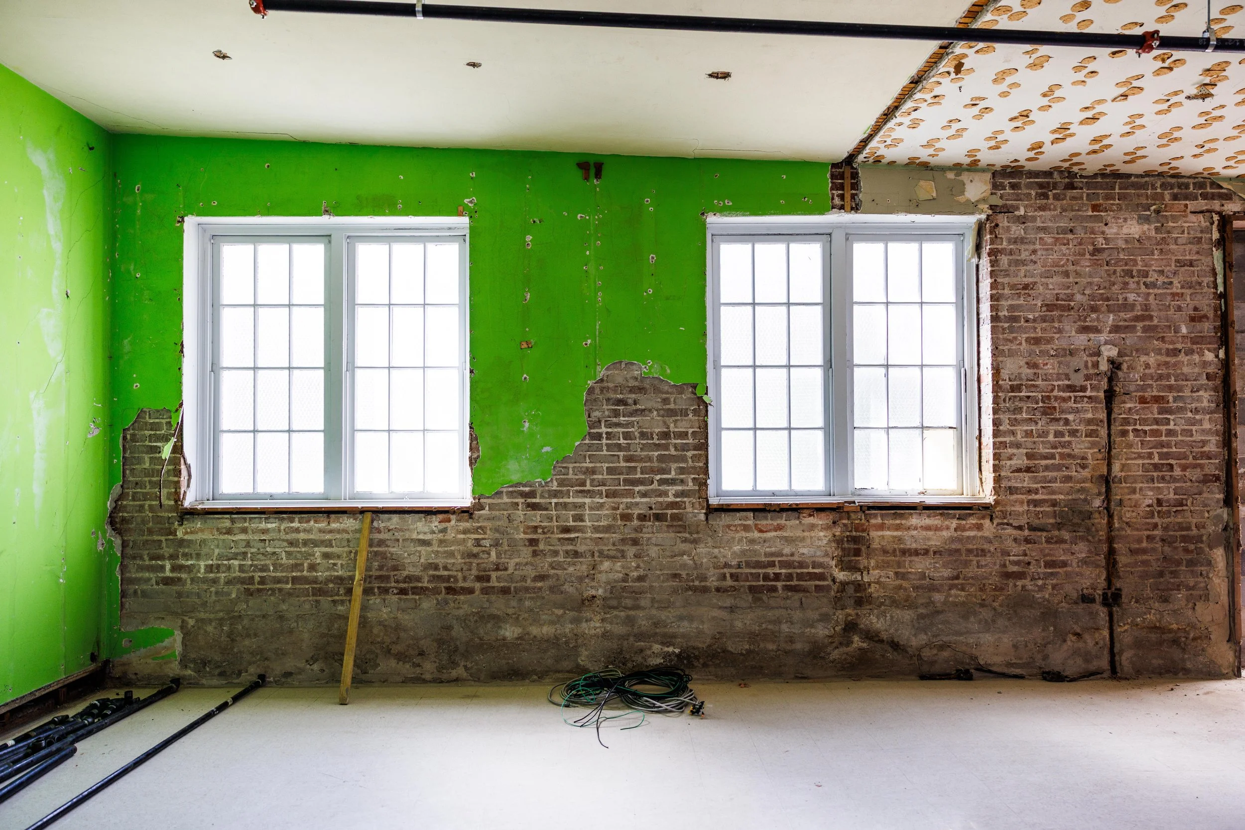 Interior room under renovation with exposed brick wall and two white-framed windows. The wall has patches of green paint and partially removed drywall, revealing bricks. Construction materials and electrical wiring on the floor.
