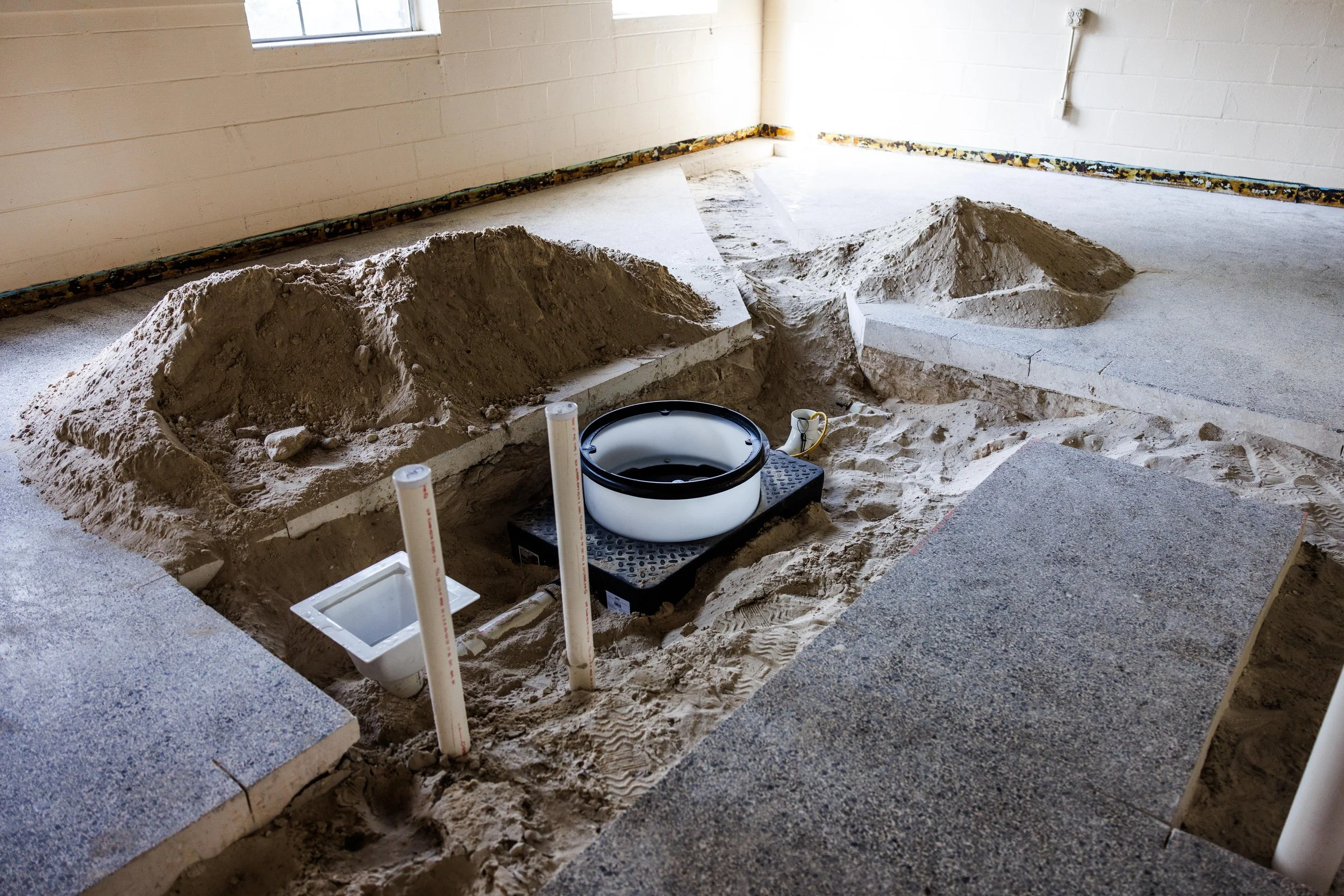 A construction site indoor with a partially dug hole in the floor, measuring tools, and a small white bathroom fixture, possibly a toilet, in the hole.