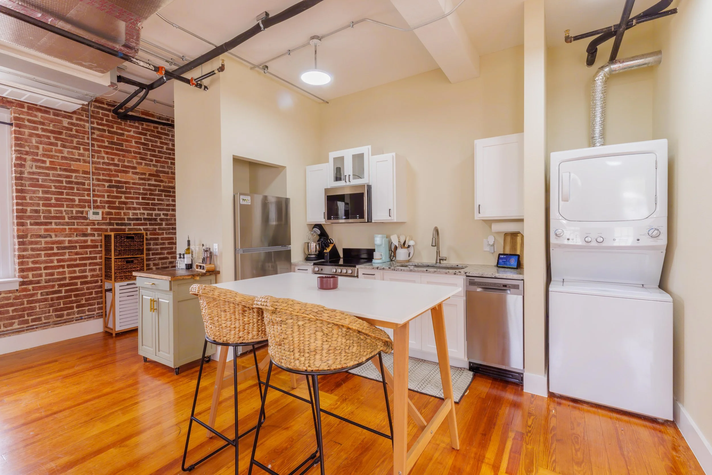 Open kitchen with white cabinets, stainless steel refrigerator, microwave, and dishwasher, a small island with two wicker chairs, brick accent wall, and a stacked washer and dryer.