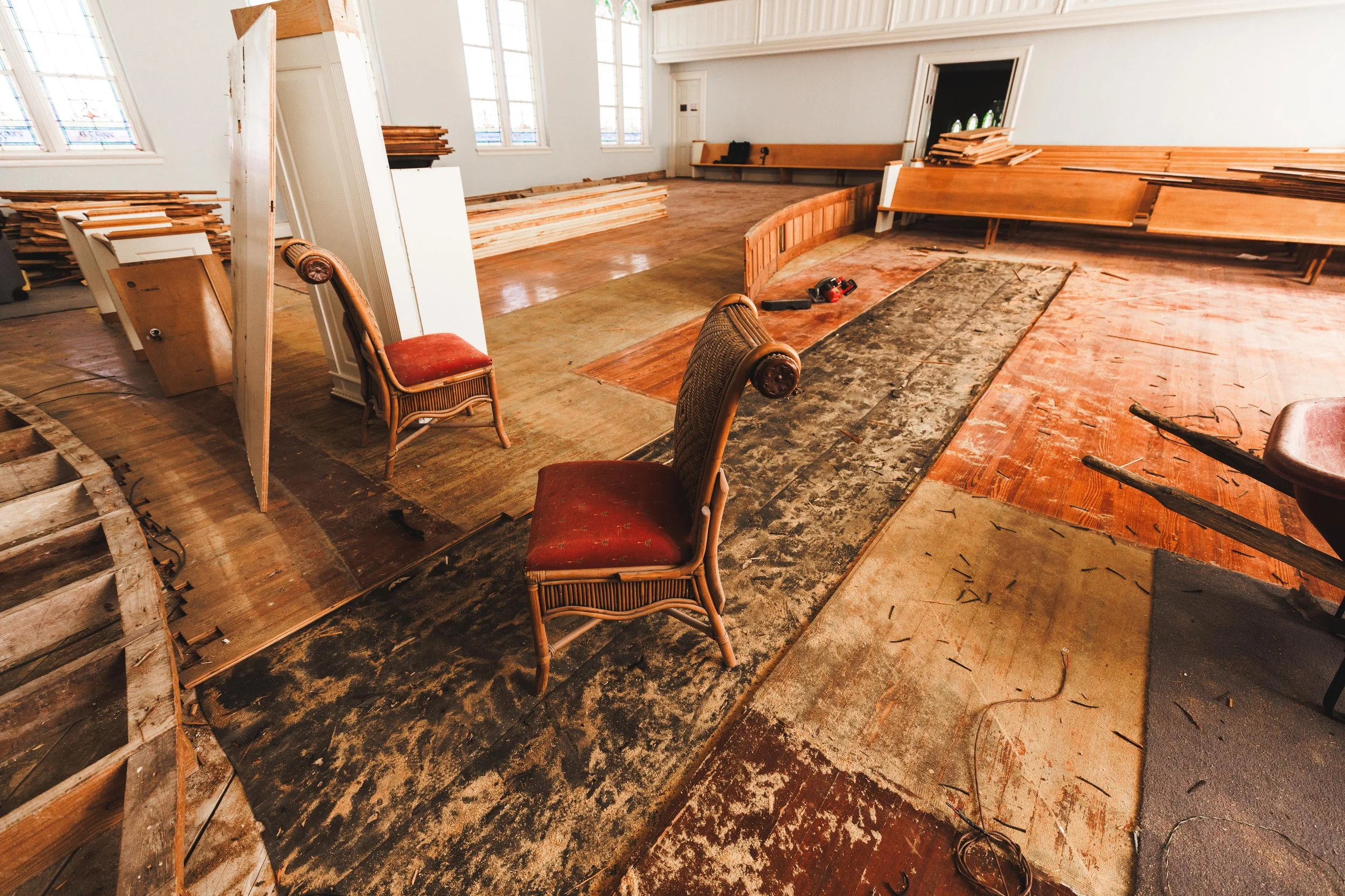 Interior of a space undergoing renovation with broken and removed flooring, wooden chairs, and construction tools scattered around.