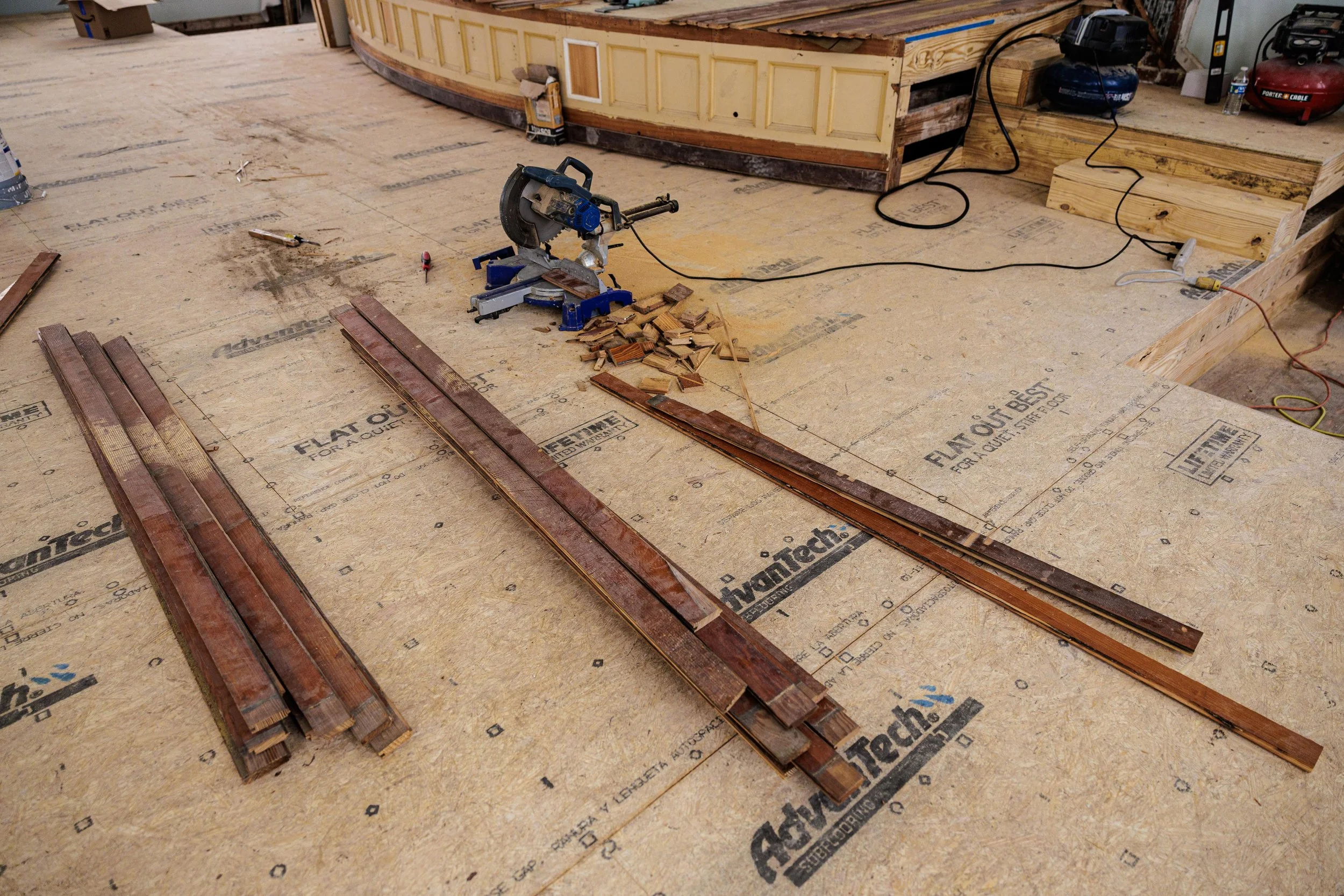 Construction site with wooden planks, tools, and equipment on a plywood floor. A miter saw with wood scraps is visible, along with a curved bar and a partially built wooden wall or cabinetry in the background.