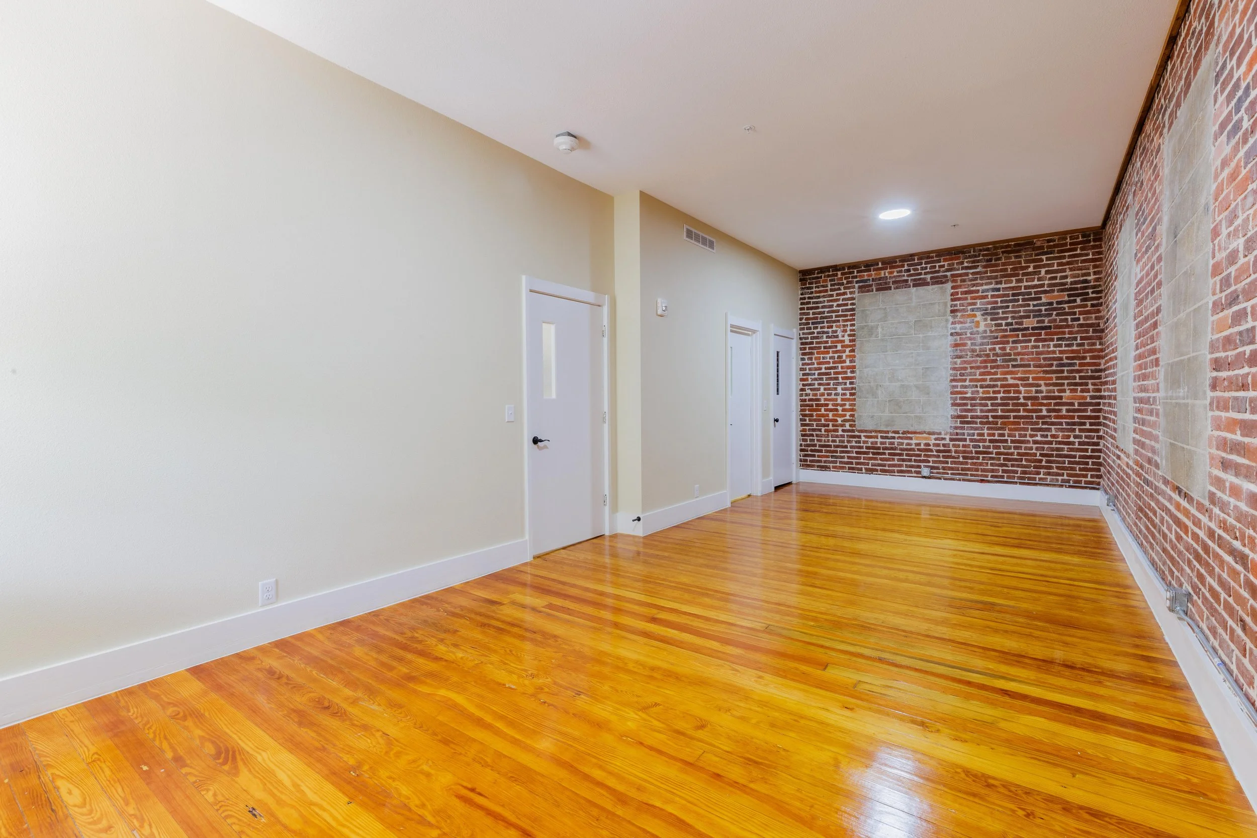 Empty room with hardwood floors, a white wall with two doors, and a brick wall with windows covered by cardboard.