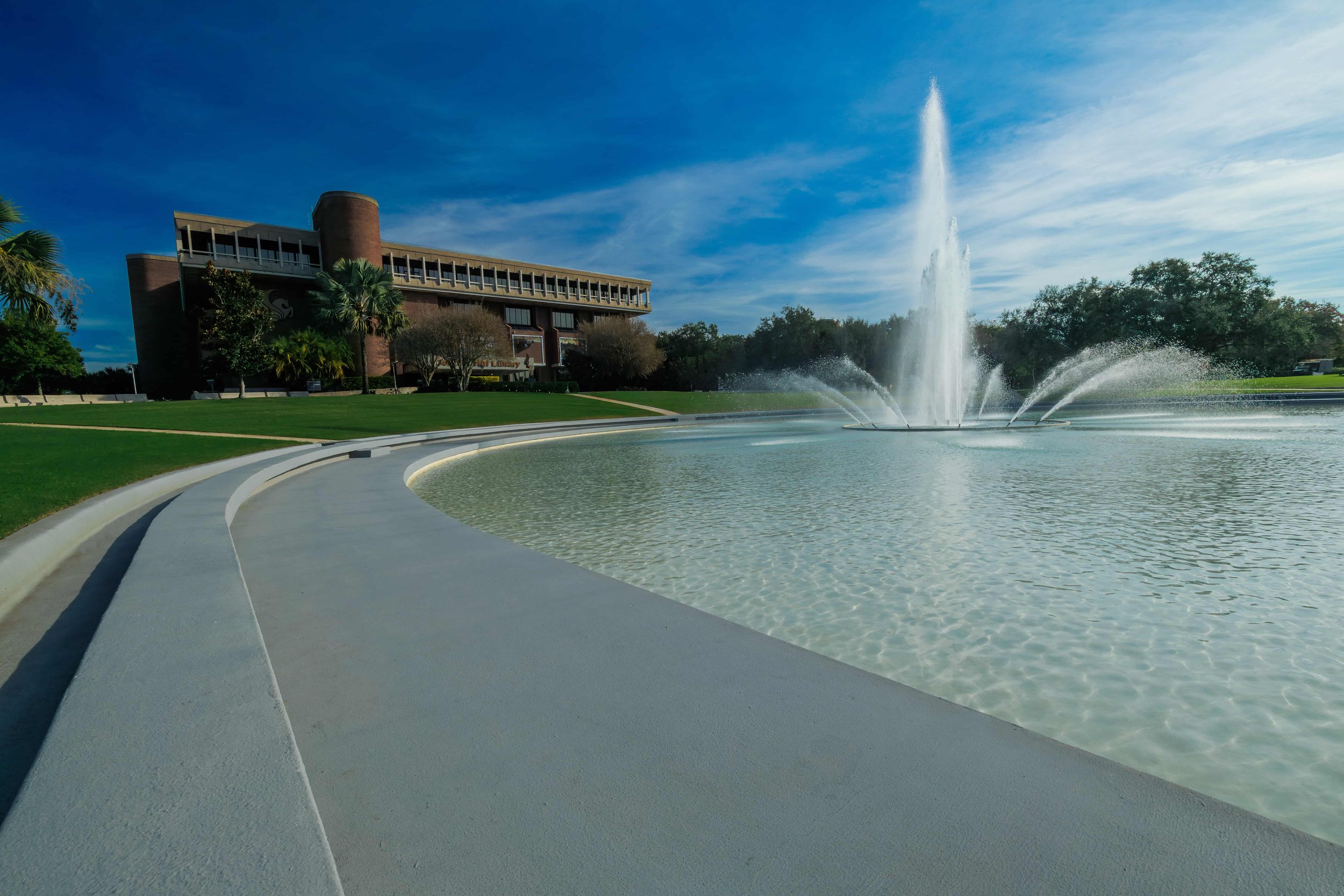 A fountain with water spraying high into the sky in a circular pool, with a modern building and green trees in the background under a blue sky.