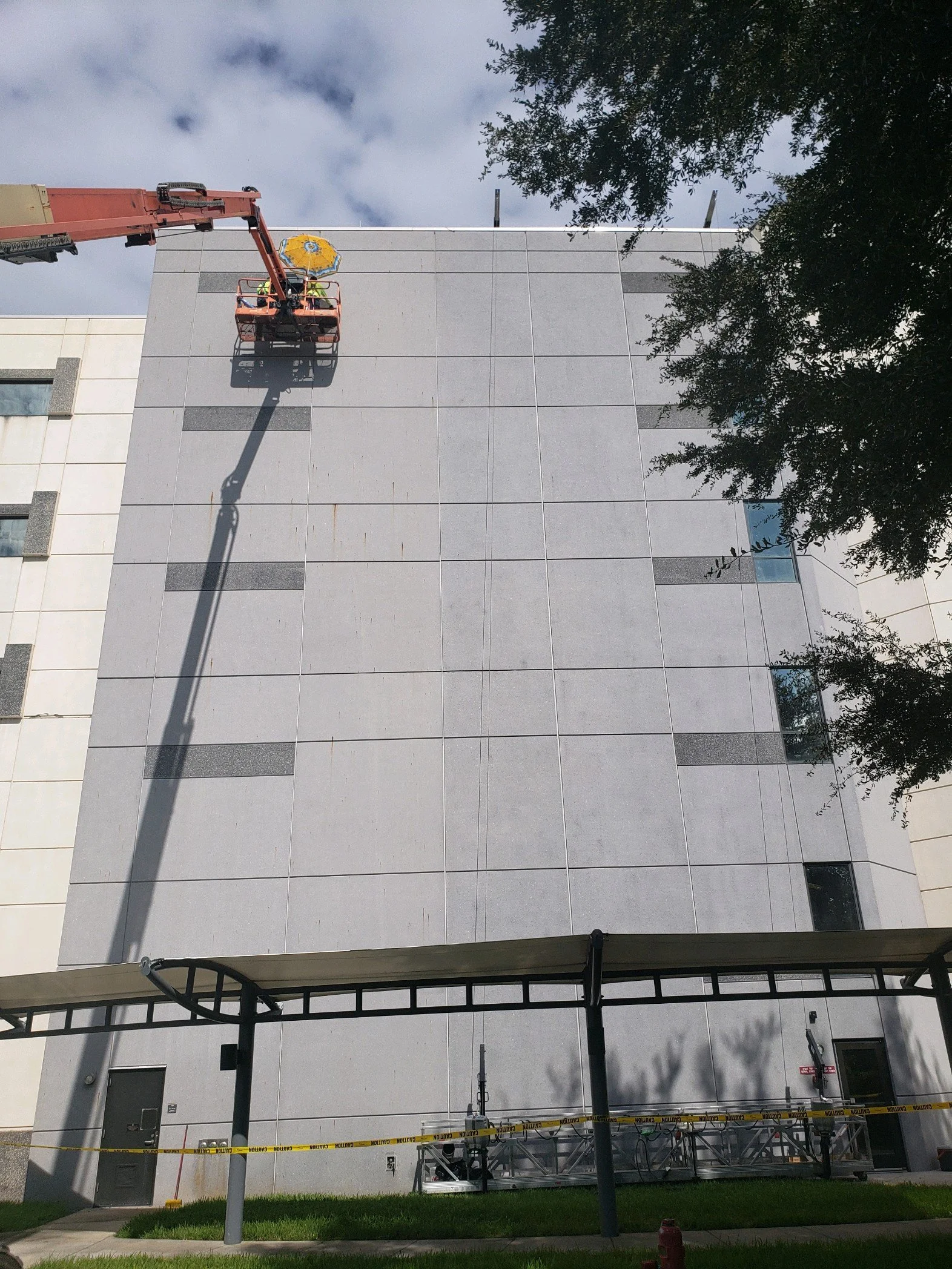 Elevated view of a building with a person working on the roof using a cherry picker, surrounded by trees, cloudy sky, and construction equipment at the ground level.
