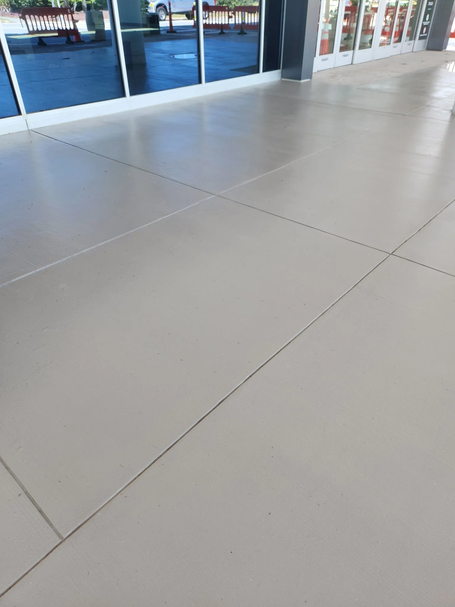 Exterior view of a building with large glass doors and windows, showing a clean sidewalk and a reflection of parked vehicles in the glass.