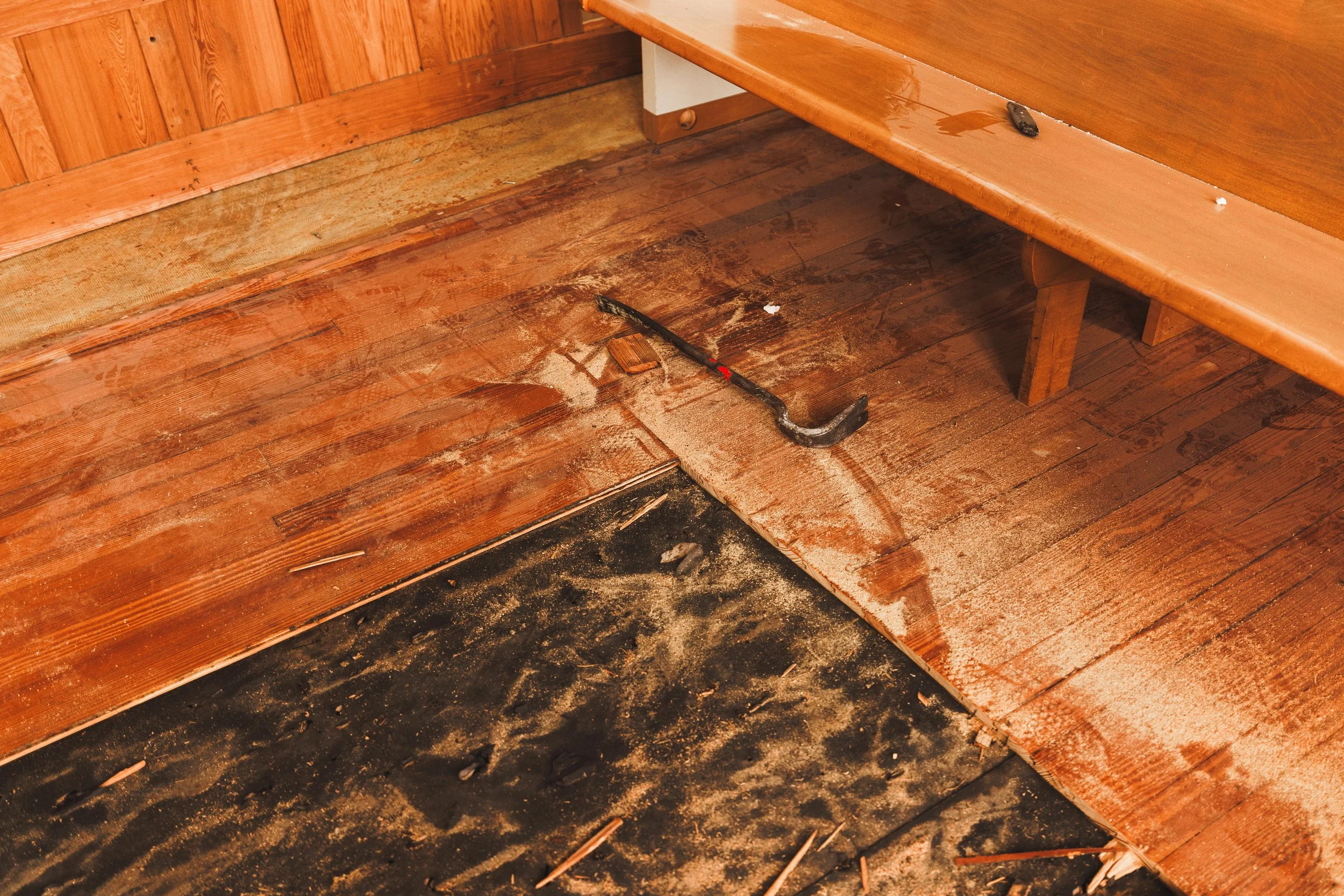 The image shows a damaged wooden floor with sawdust, an angled crowbar, part of a wooden bench, and some scattered wood chips and nails.