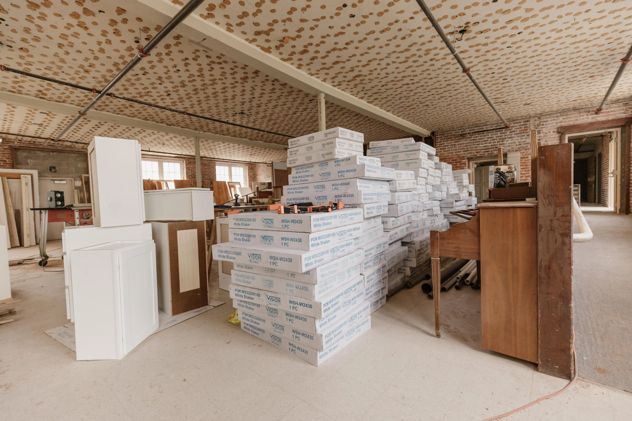 A storage room with stacks of white boxes labeled 'White Shaker' and kitchen cabinets, pipes, and a piano.