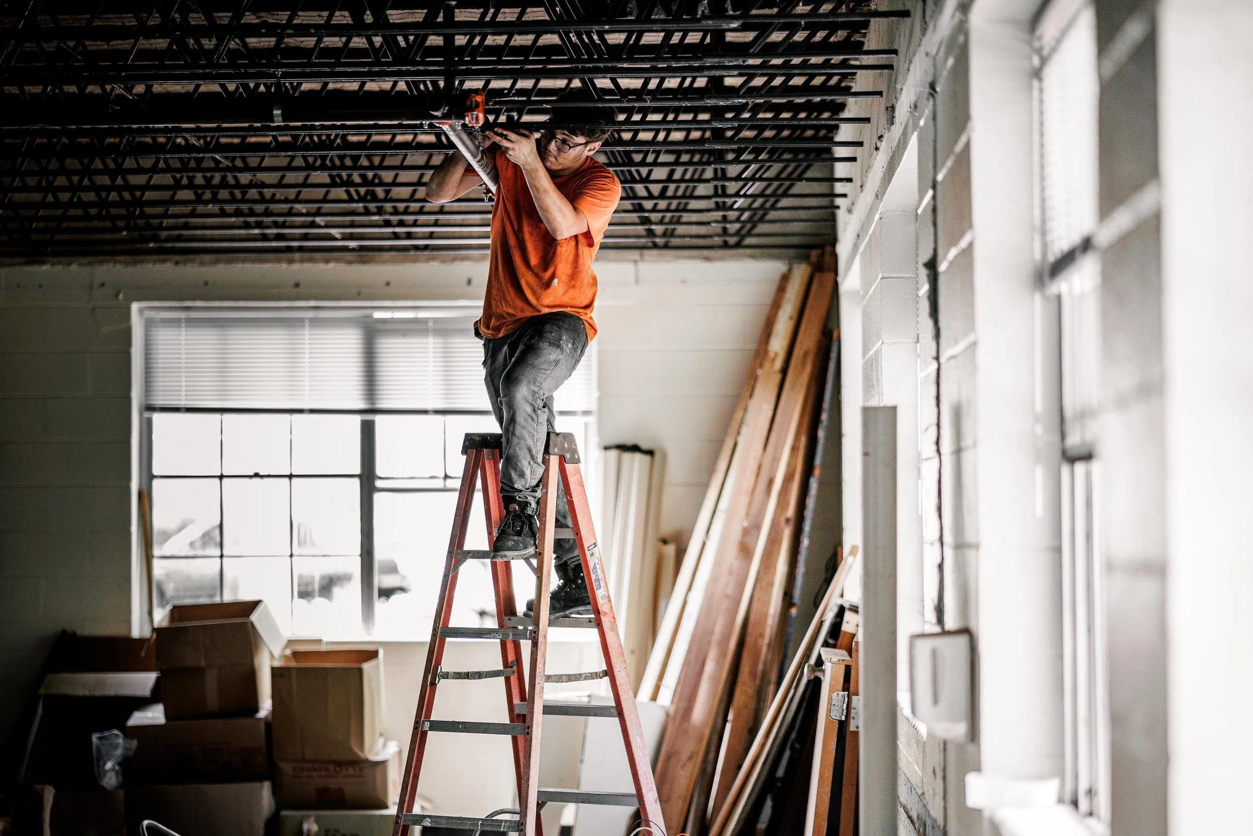 A man standing on a ladder working on the ceiling in a room with construction materials and a large window.