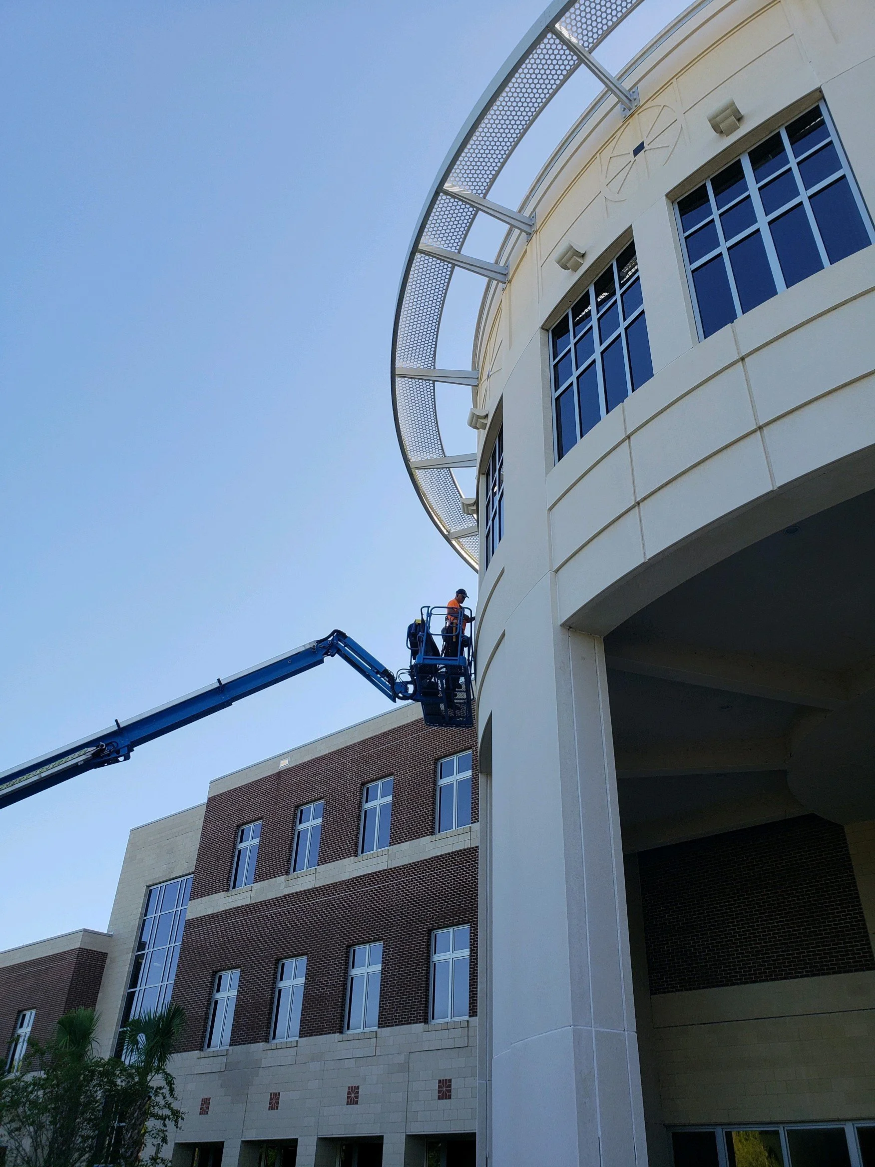 A worker in an orange safety vest and helmet operating a crane lift outside a large, modern building, possibly performing maintenance or construction work.