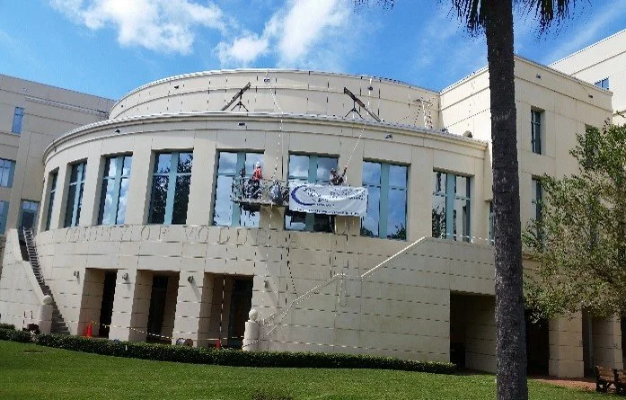 Construction workers on a suspended scaffold working on the large front windows of an art deco style beige building with a curved facade, palm tree, and blue sky in the background.