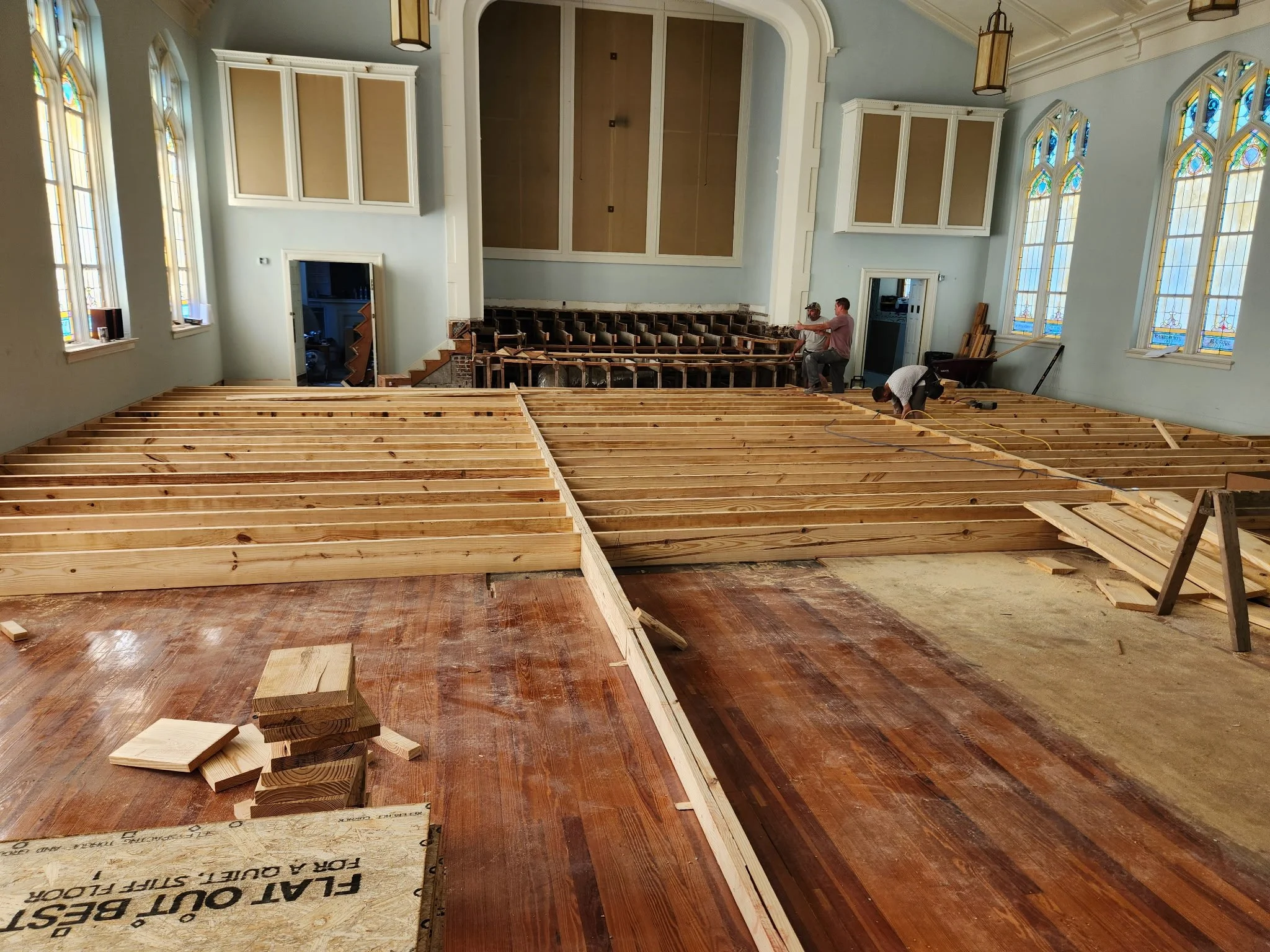 Inside a church undergoing renovation, workers install new wooden flooring with visible wooden beams, stained glass windows, and a high ceiling with hanging lanterns.