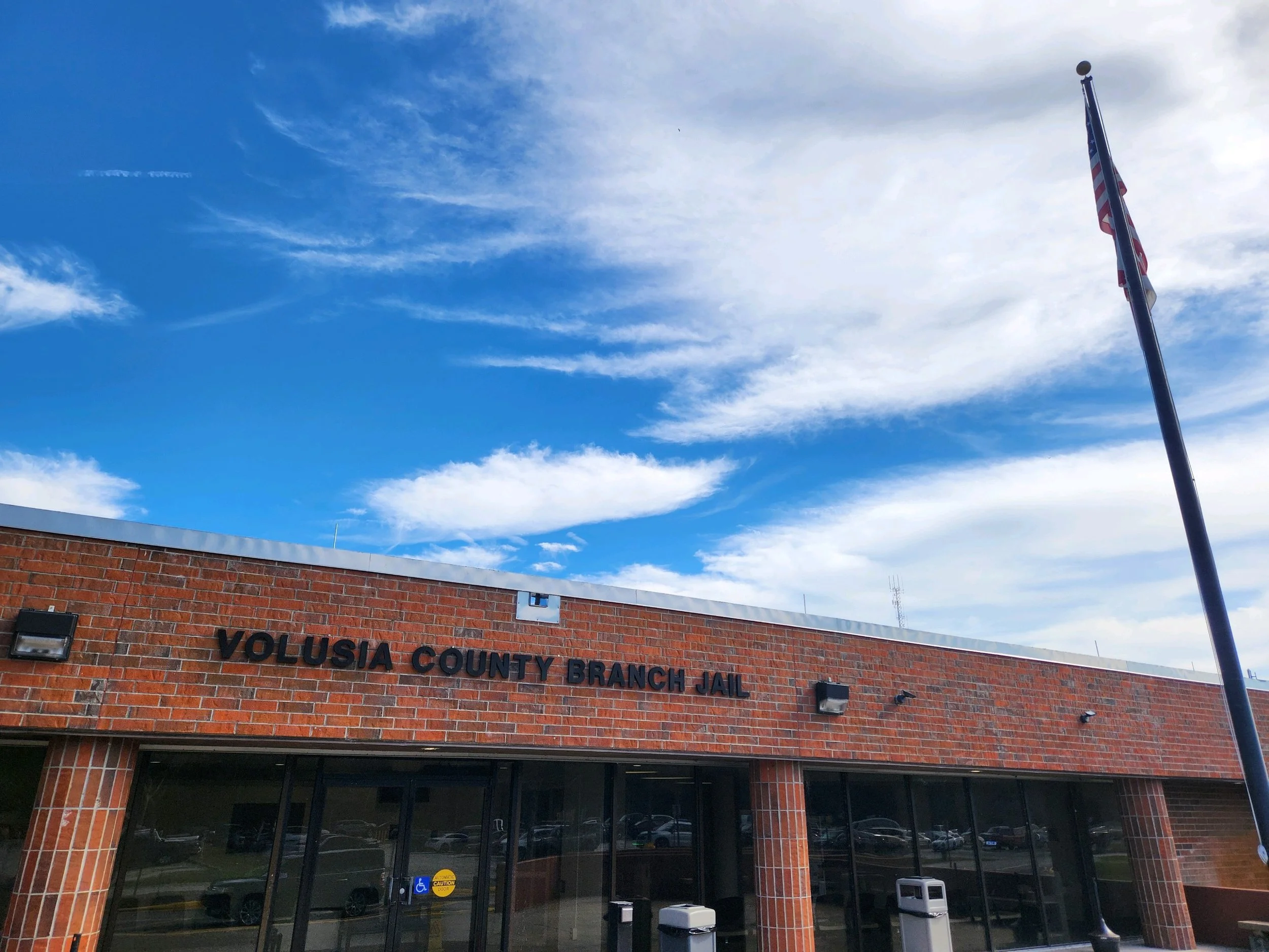 The exterior of Volusia County Branch Jail building made of red brick, with a sign displaying the name, under a partly cloudy blue sky, with an American flag on a pole to the right.