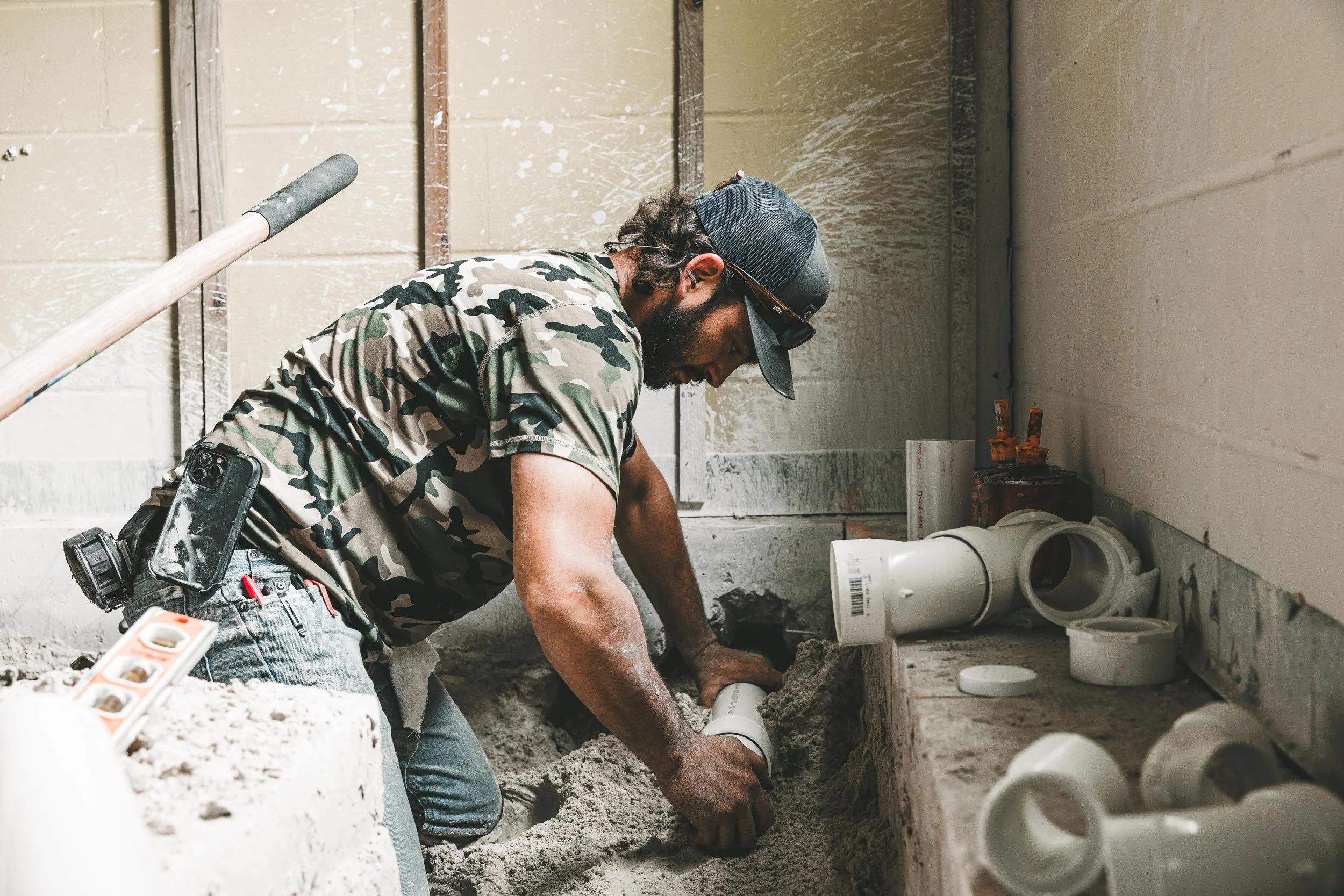 A man working on plumbing inside a construction site, wearing a camouflage shirt and a black cap, installing pipes.