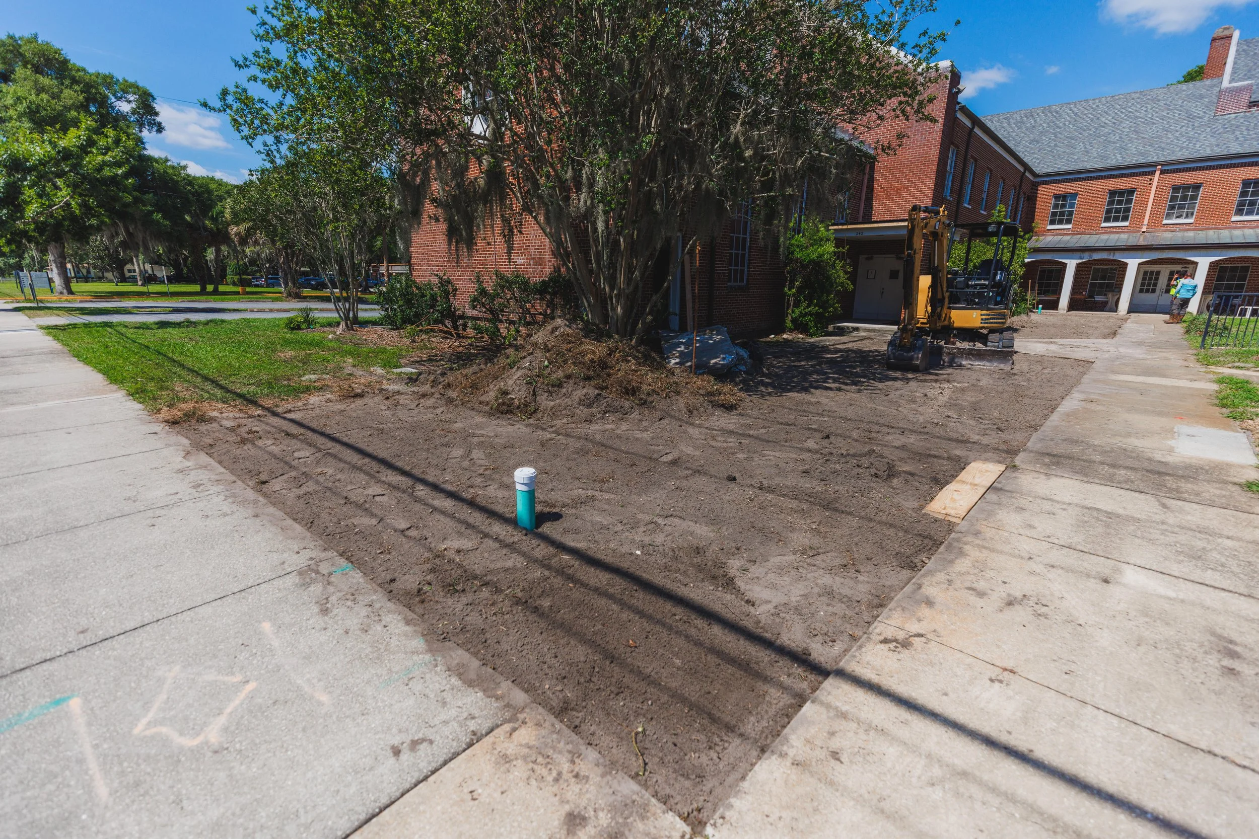 A construction site on a sidewalk, with dirt and a small excavator, beside a brick building and trees, under a blue sky.
