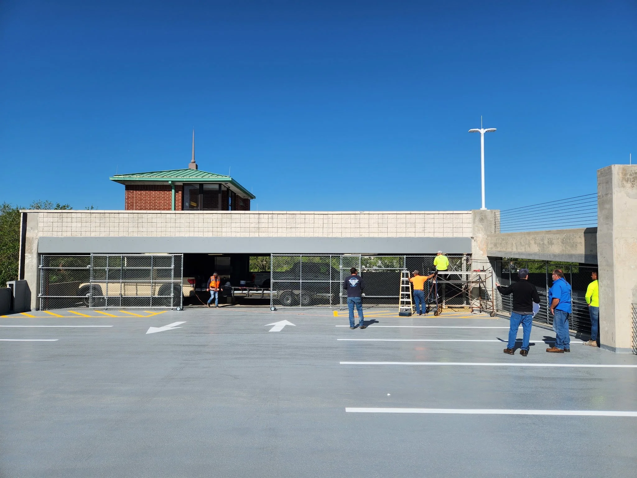 Construction workers installing security fencing at the entrance of a parking garage on a clear, sunny day with a blue sky.