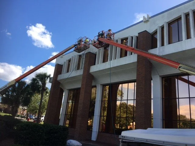 Workers on lift doing maintenance work on the exterior of a multi-story building with large windows, clear blue sky and palm trees in the background.