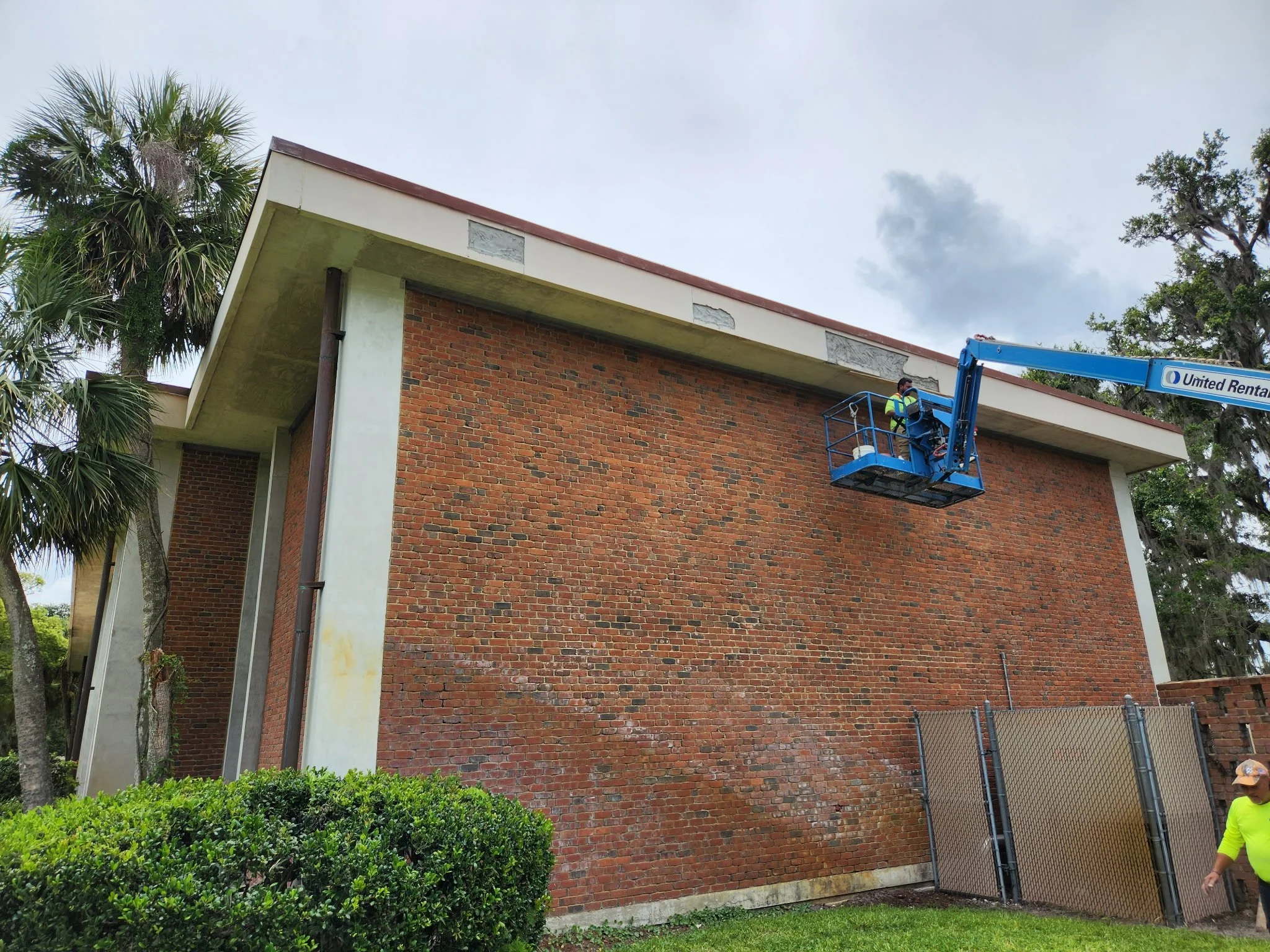 A worker on a blue lift inspecting a brick building's exterior wall that is under repair, with cloudy sky overhead and greenery at the base.