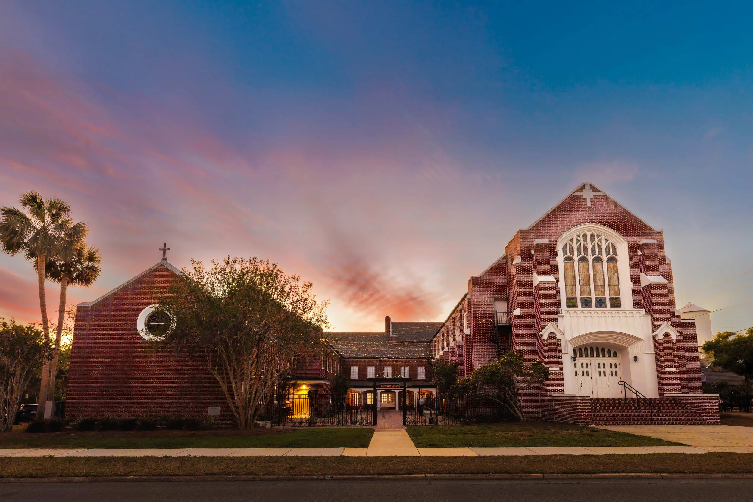 A brick church with gothic windows and a cross on top, set against a colorful sunset sky, with palm trees on the left and a sidewalk in front.